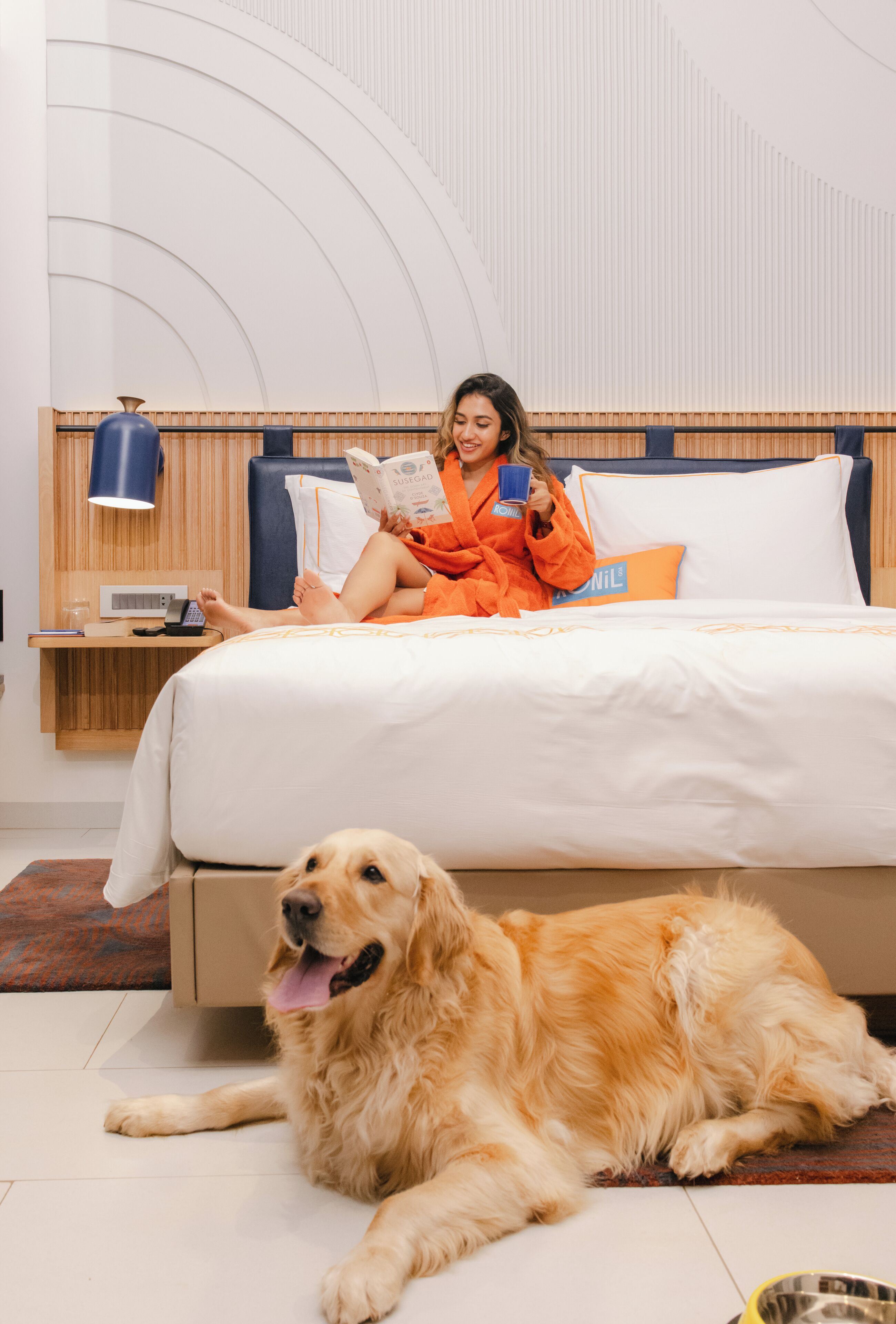 Une personne est assise sur un lit, en train de lire un livre, et un chien est allongé à ses côtés.
