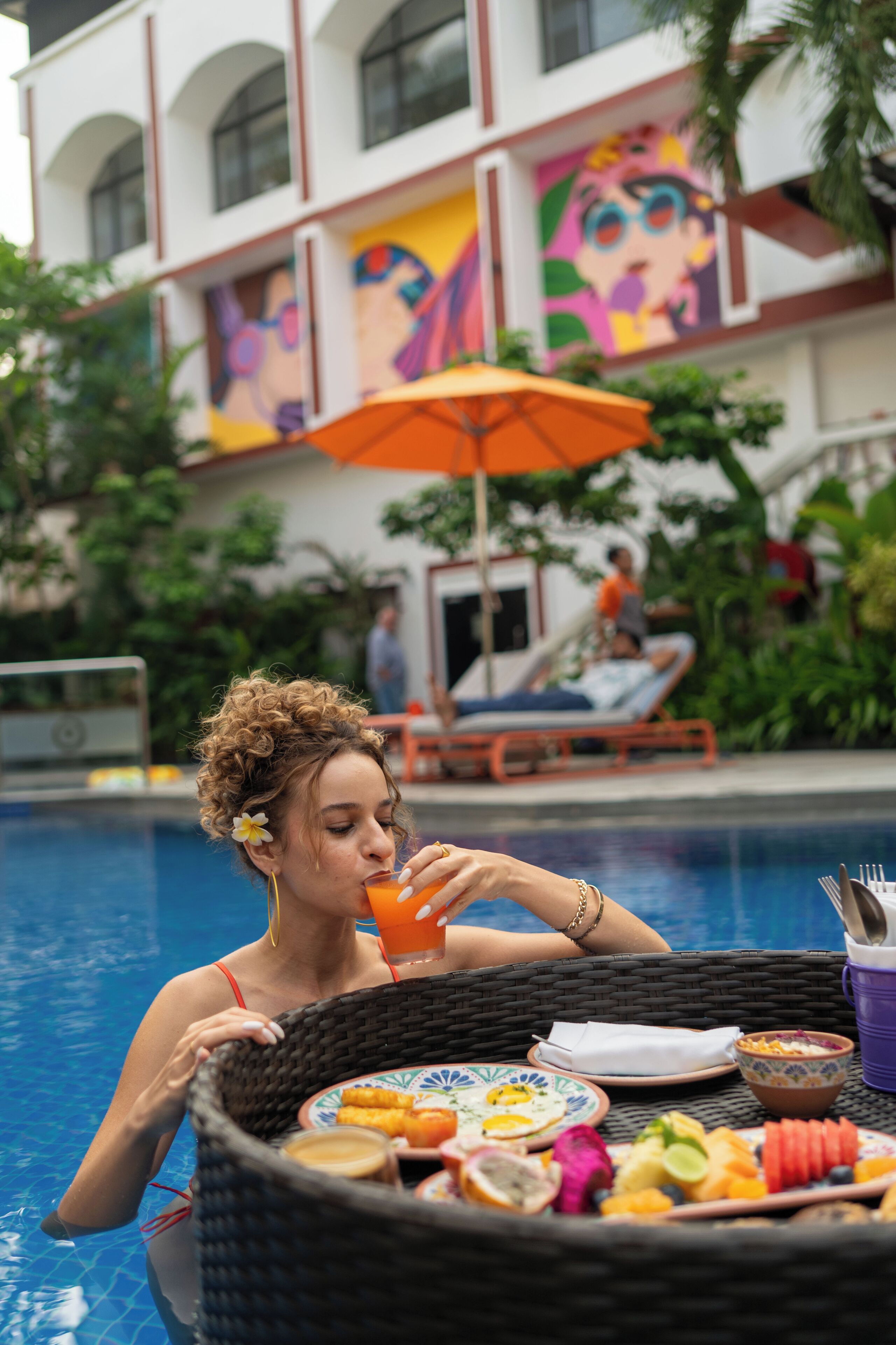 A poolside dining area with a wicker table set with food and drinks, a colorful mural on the building, and a person relaxing by the pool.