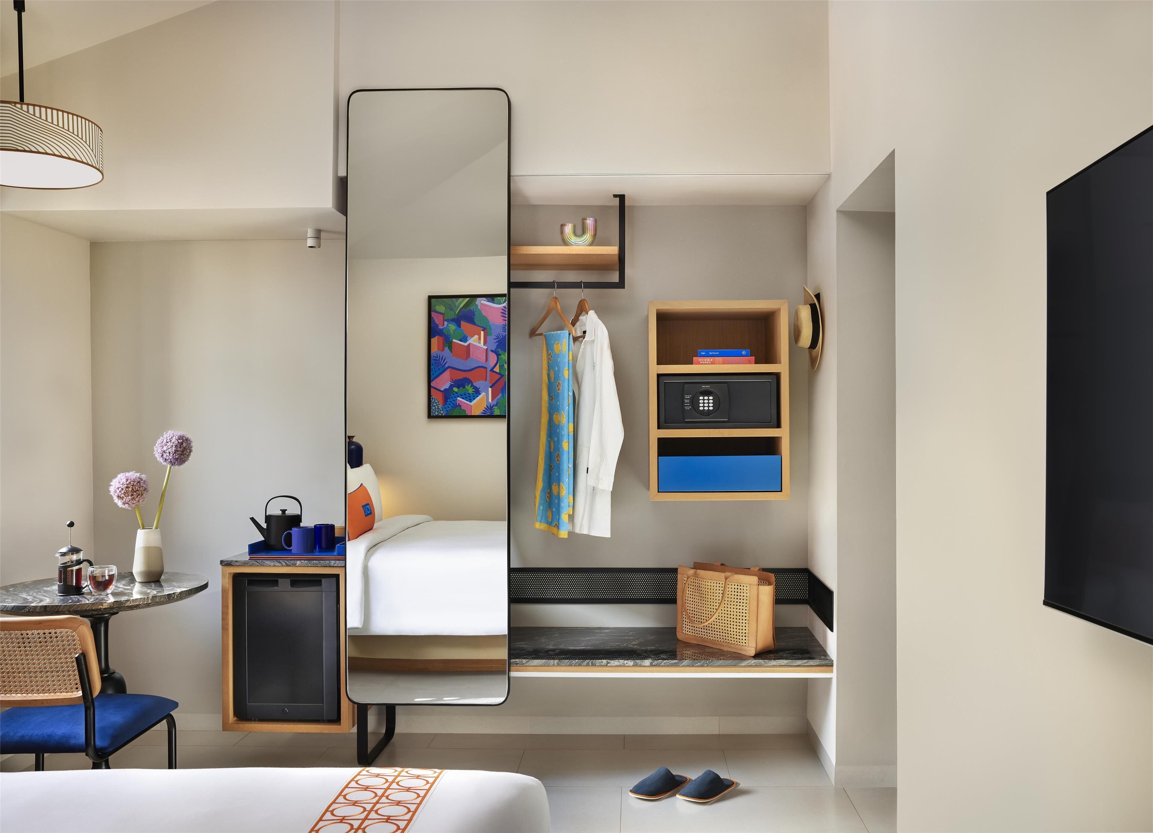 A modern hotel room with a bed, a small table with a vase and cups, a chair, a mirror, a shelf with books and a hat, and a bench with slippers.