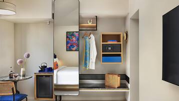 A modern hotel room with a bed, a small table with a vase and cups, a chair, a mirror, a shelf with books and a hat, and a bench with slippers.