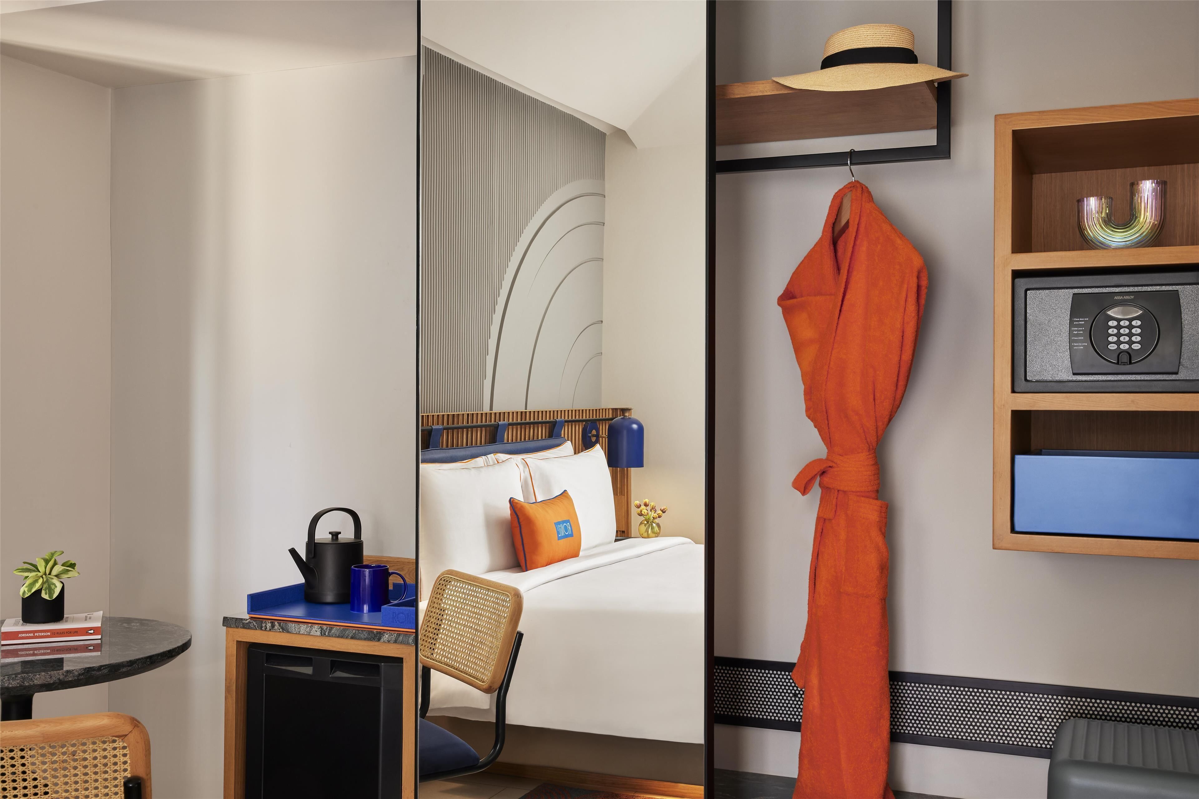 A hotel room with a bed, a desk with a kettle, a chair, a small table with a plant, and a robe hanging on a hook.