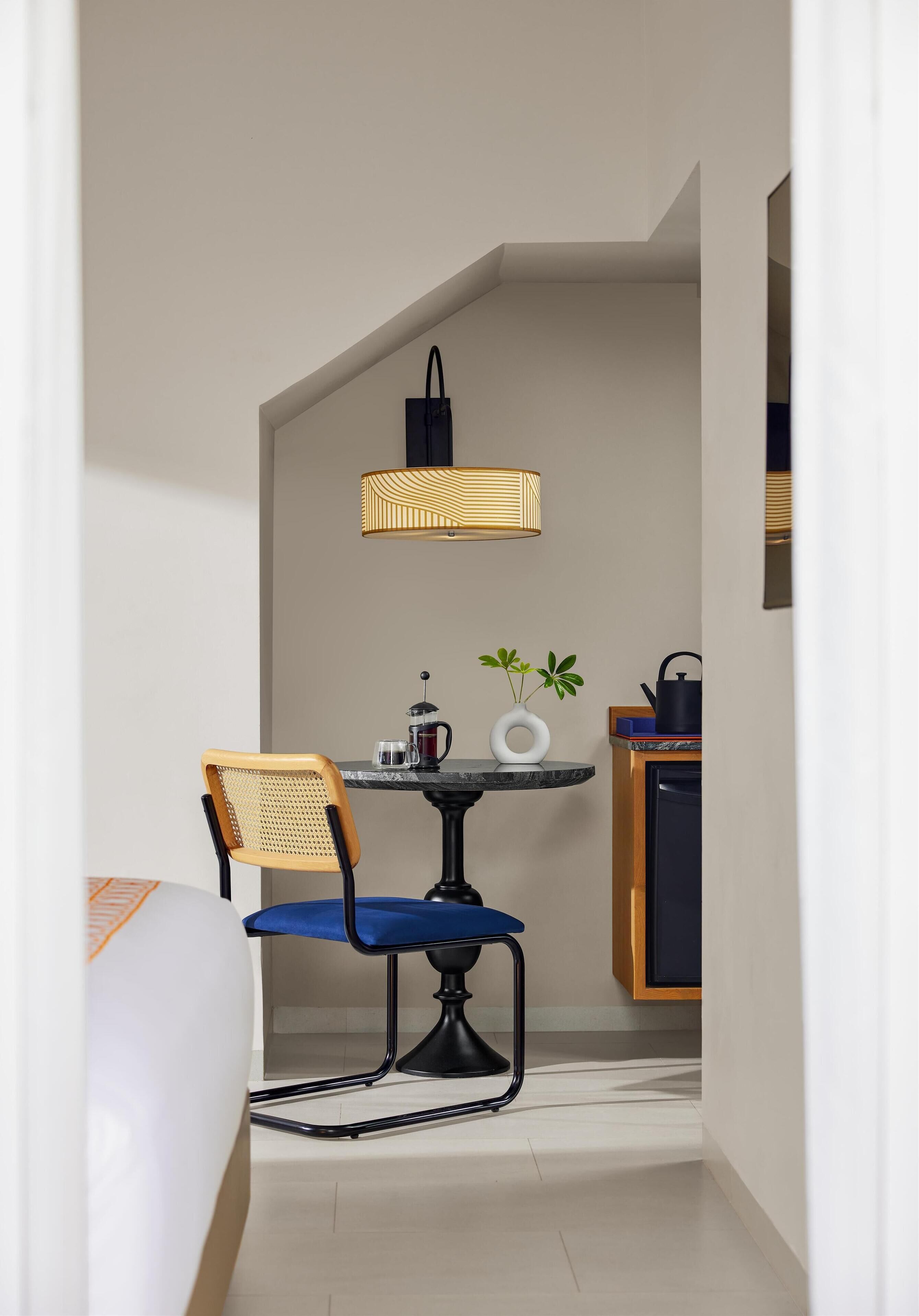A compact hotel room with a dining area featuring a small table, a chair, a lamp, a plant, and a kettle.