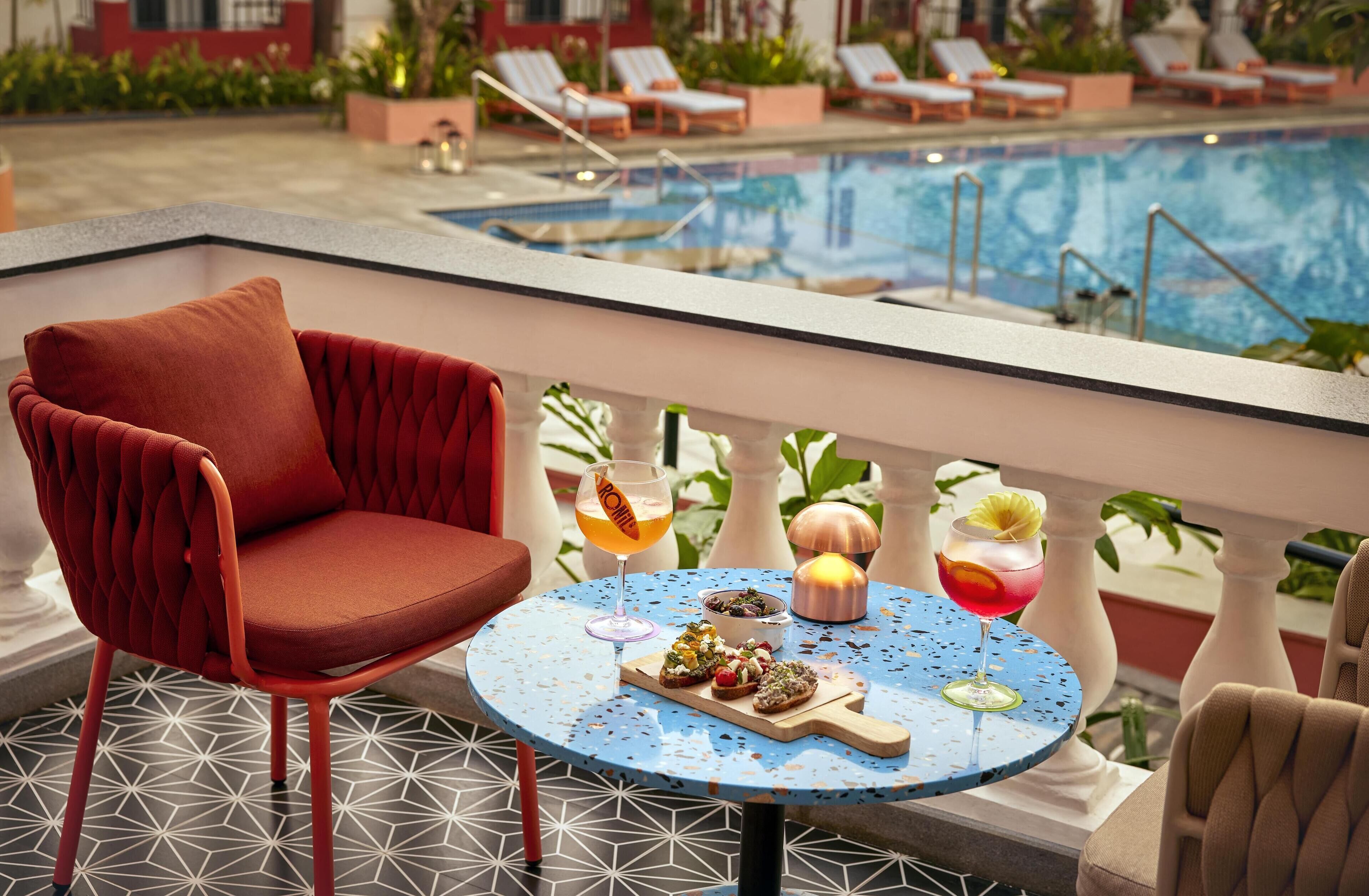 Un balcón con una silla roja, una mesa redonda con comida y bebidas, y una piscina al fondo.