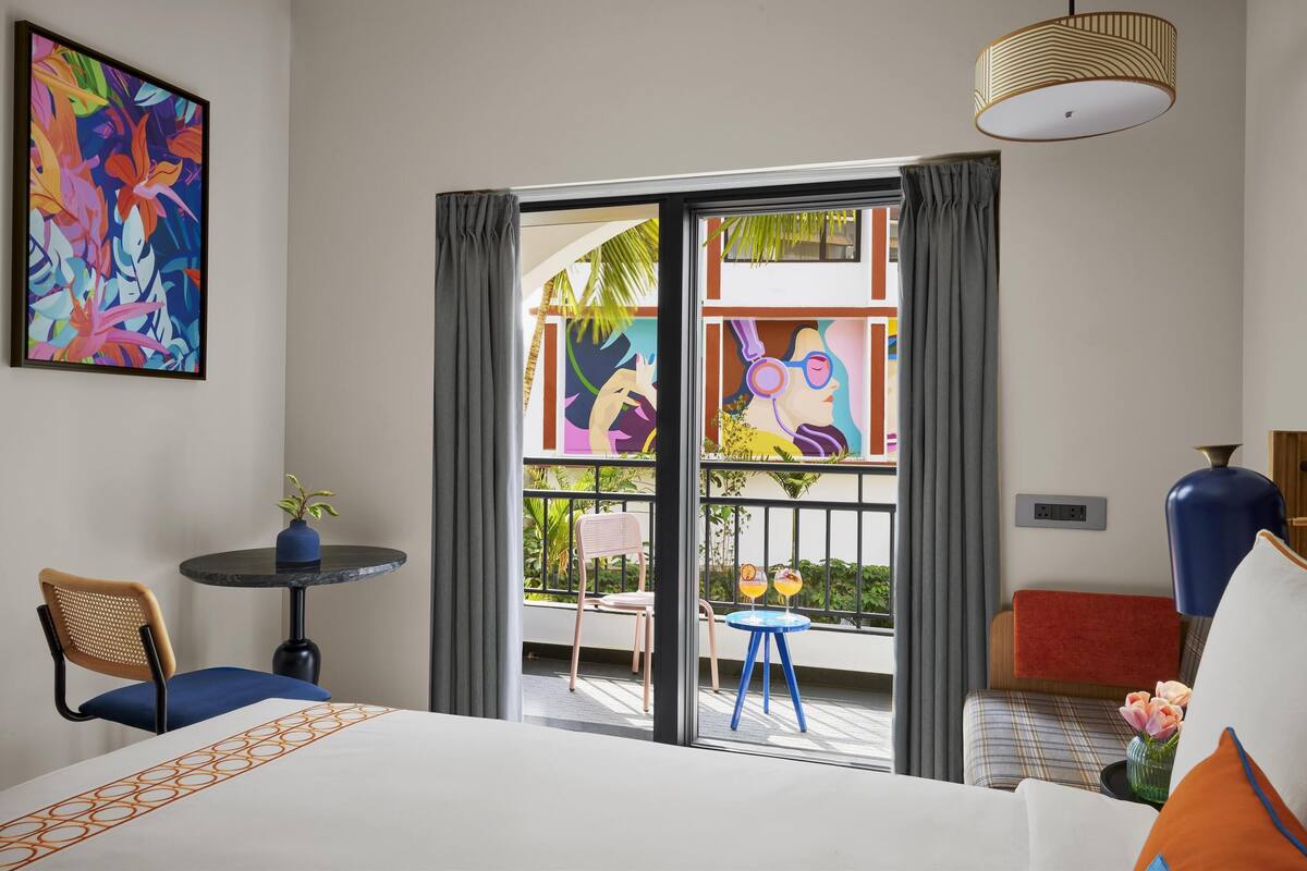 Une chambre d’hôtel avec un lit, une petite table ornée d’une plante, deux chaises, un balcon donnant sur l’extérieur et un tableau coloré accroché au mur.