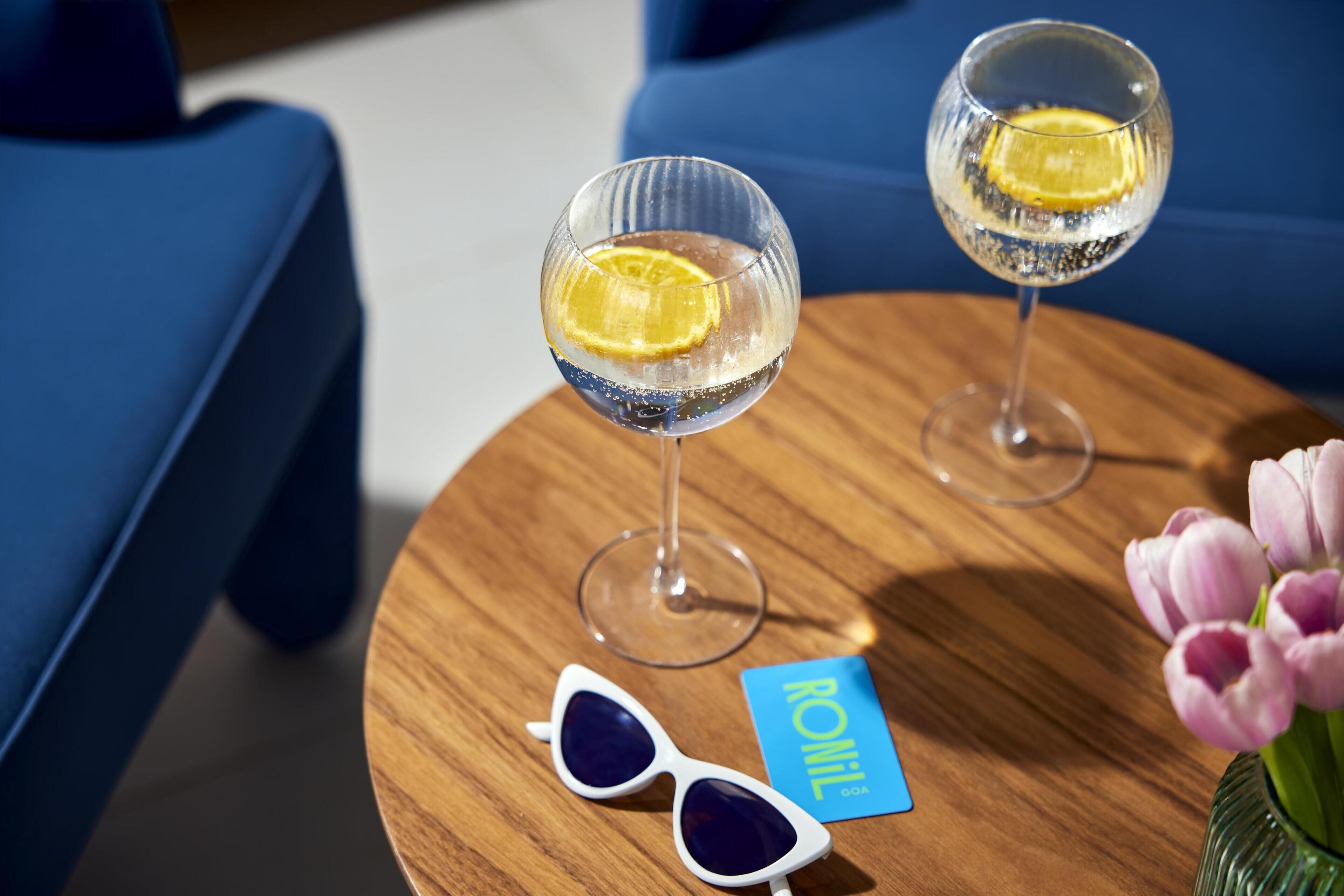 Two glasses of a clear beverage with a slice of lemon on a wooden table, a pair of sunglasses, a blue card, and a vase with flowers.
