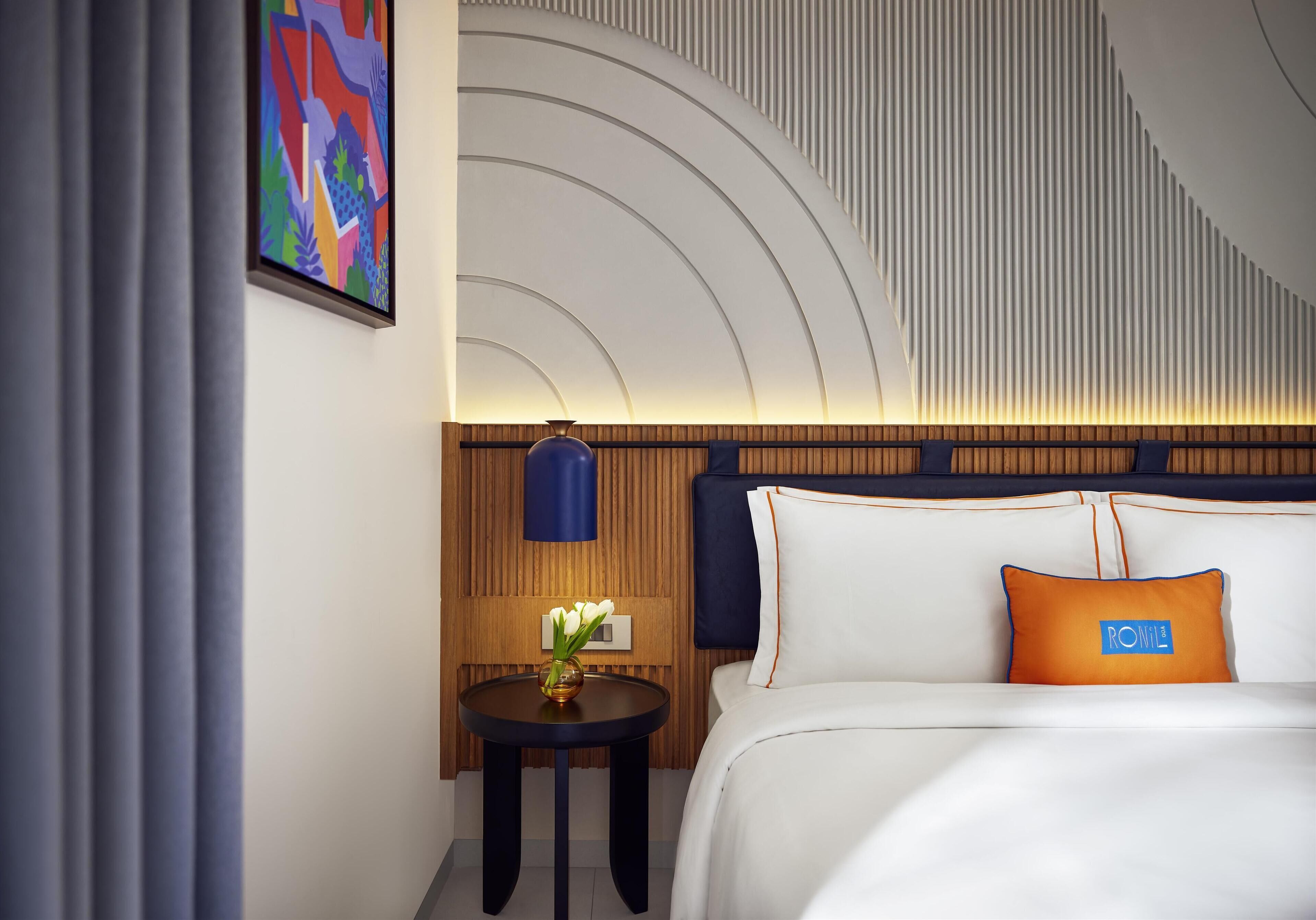 A hotel room with a bed, a bedside table with a vase of flowers, a wall art piece, and a unique ceiling design.