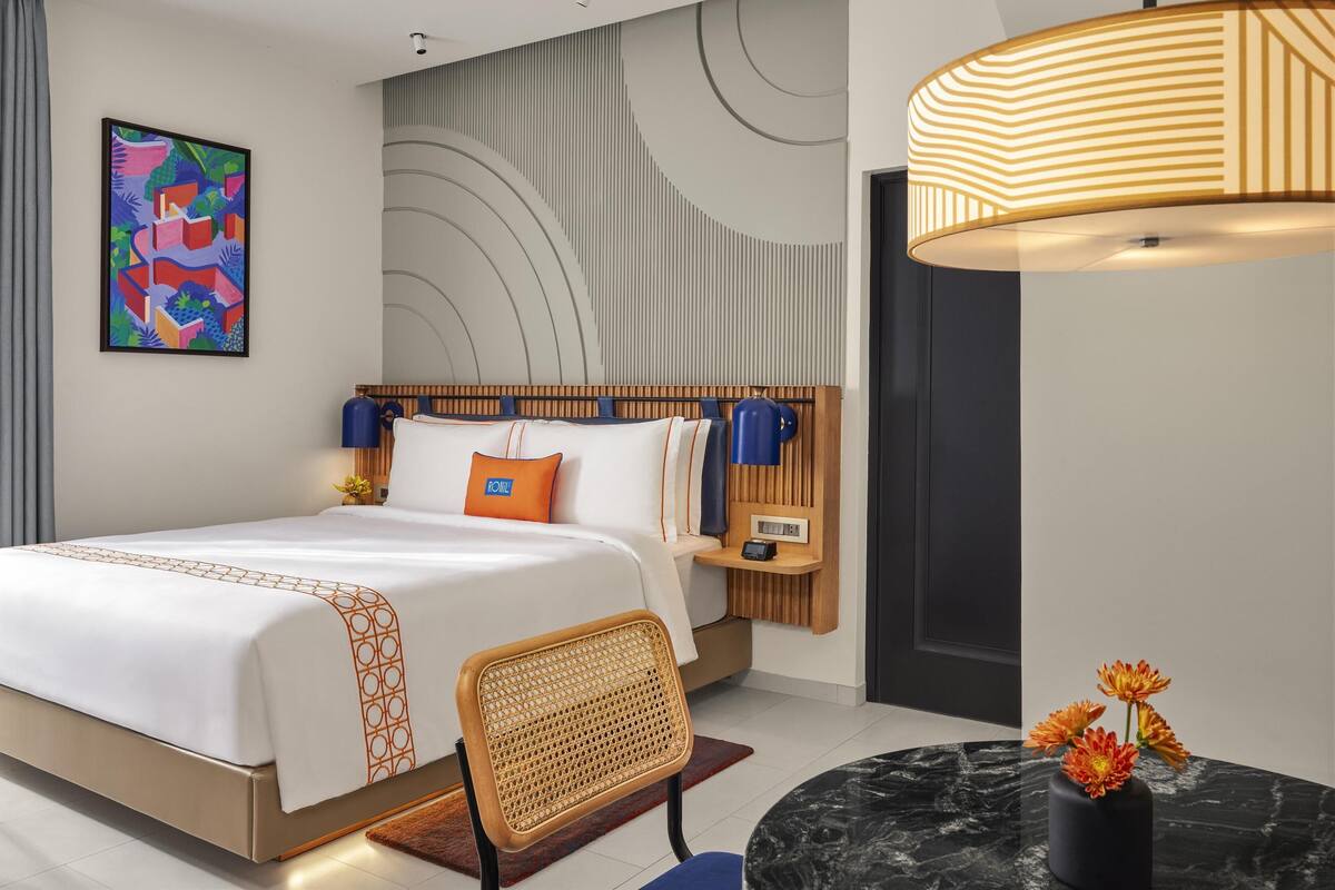 Une chambre d’hôtel moderne avec un grand lit, une table de chevet, une chaise, un bureau, une lampe et une œuvre d’art murale.