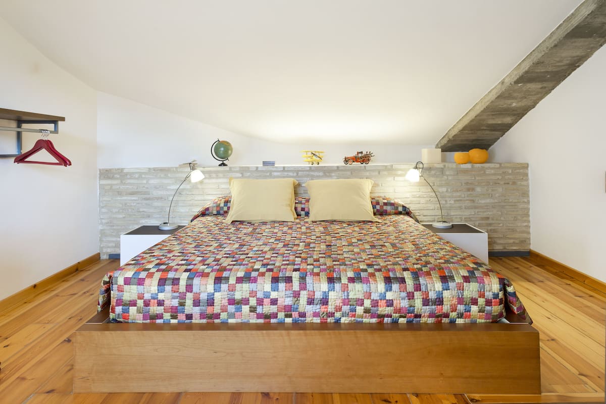 A bedroom with a wooden floor, a quilted bedspread, two bedside lamps, a brick-patterned headboard, and a red hanger on the wall.
