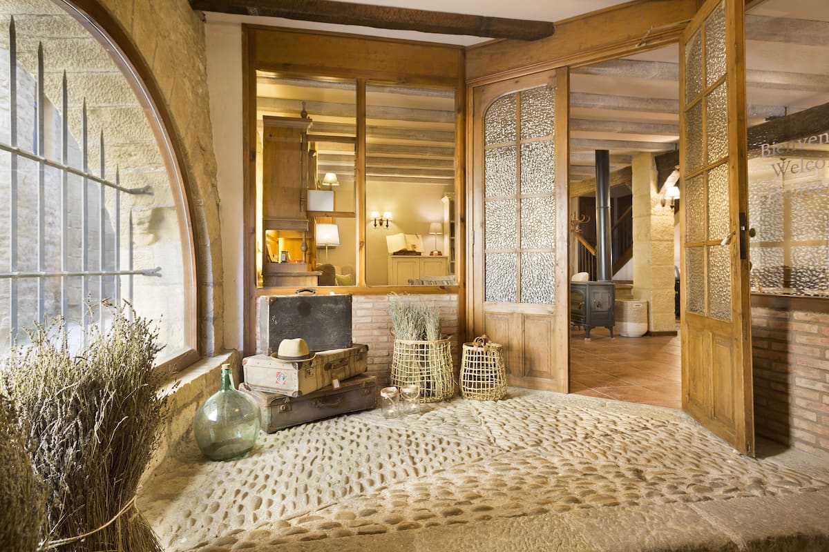 A cozy room with a stone floor, wooden beams, and a large window with metal bars. There are suitcases, a green bottle, and wicker baskets on a low table.