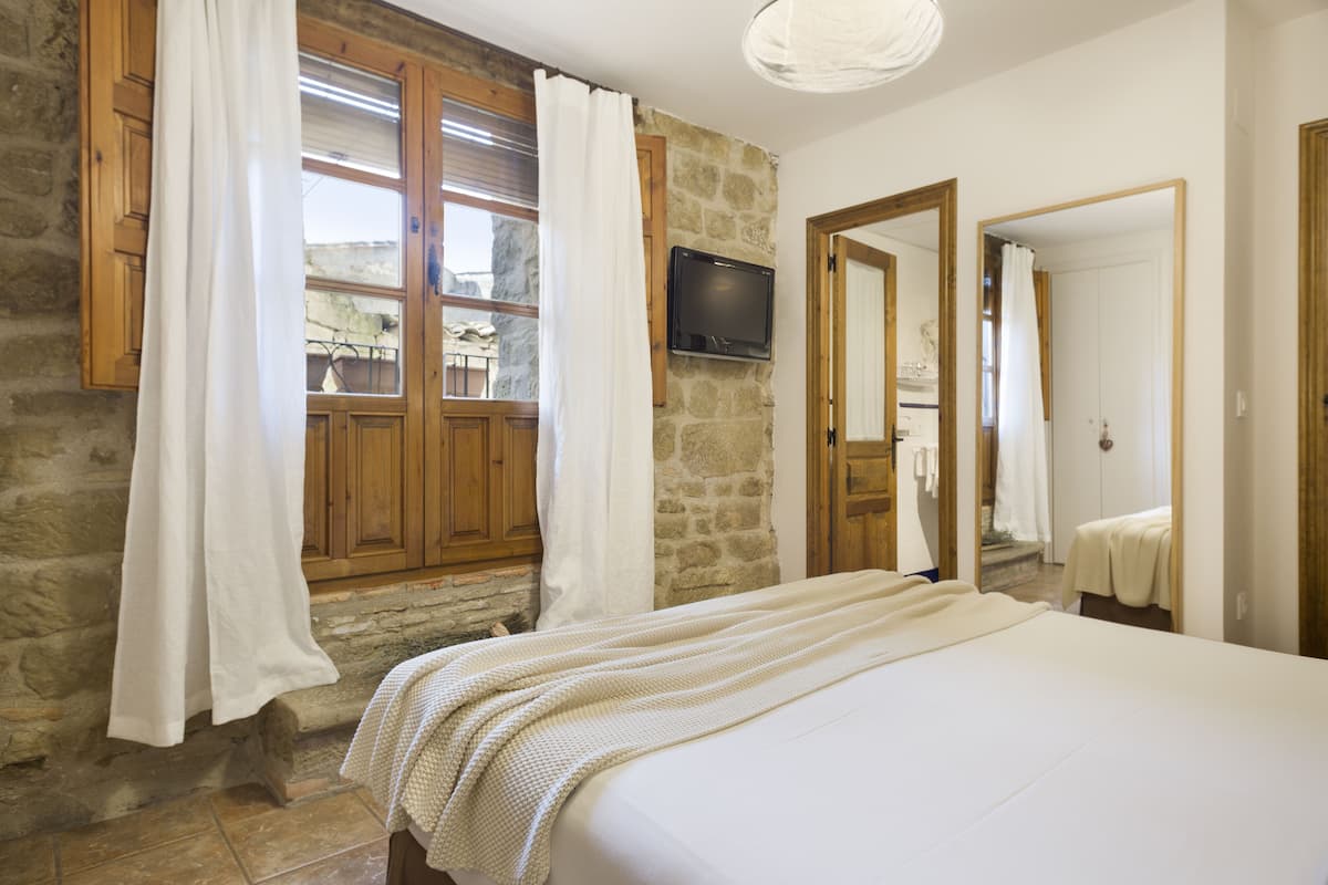 A bedroom with a stone wall, wooden doors, a bed with a white bedspread, and a TV mounted on the wall.