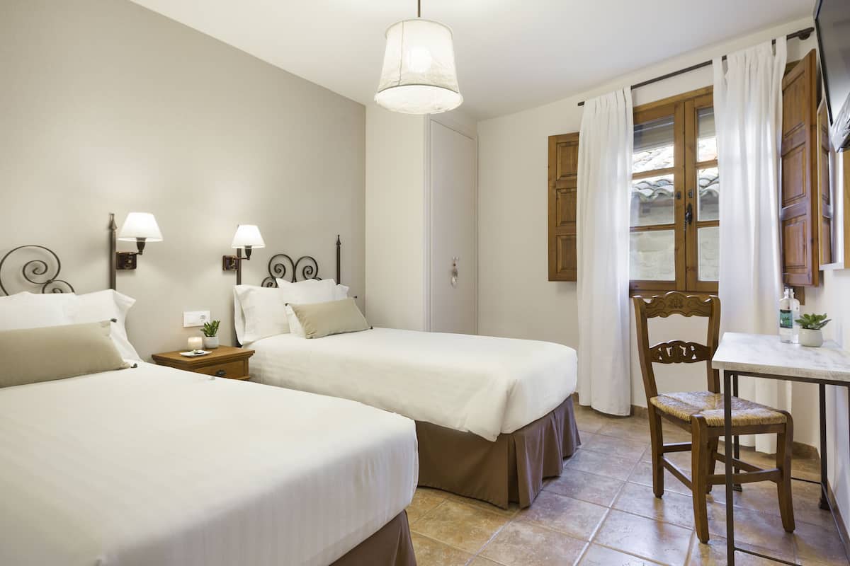 A hotel room with two beds, a wooden chair, a table, and a window with curtains.