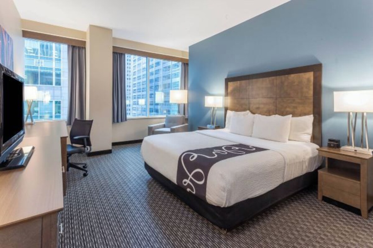 A hotel room with a large bed, a desk with a TV, two bedside lamps, and a view of a cityscape through large windows.