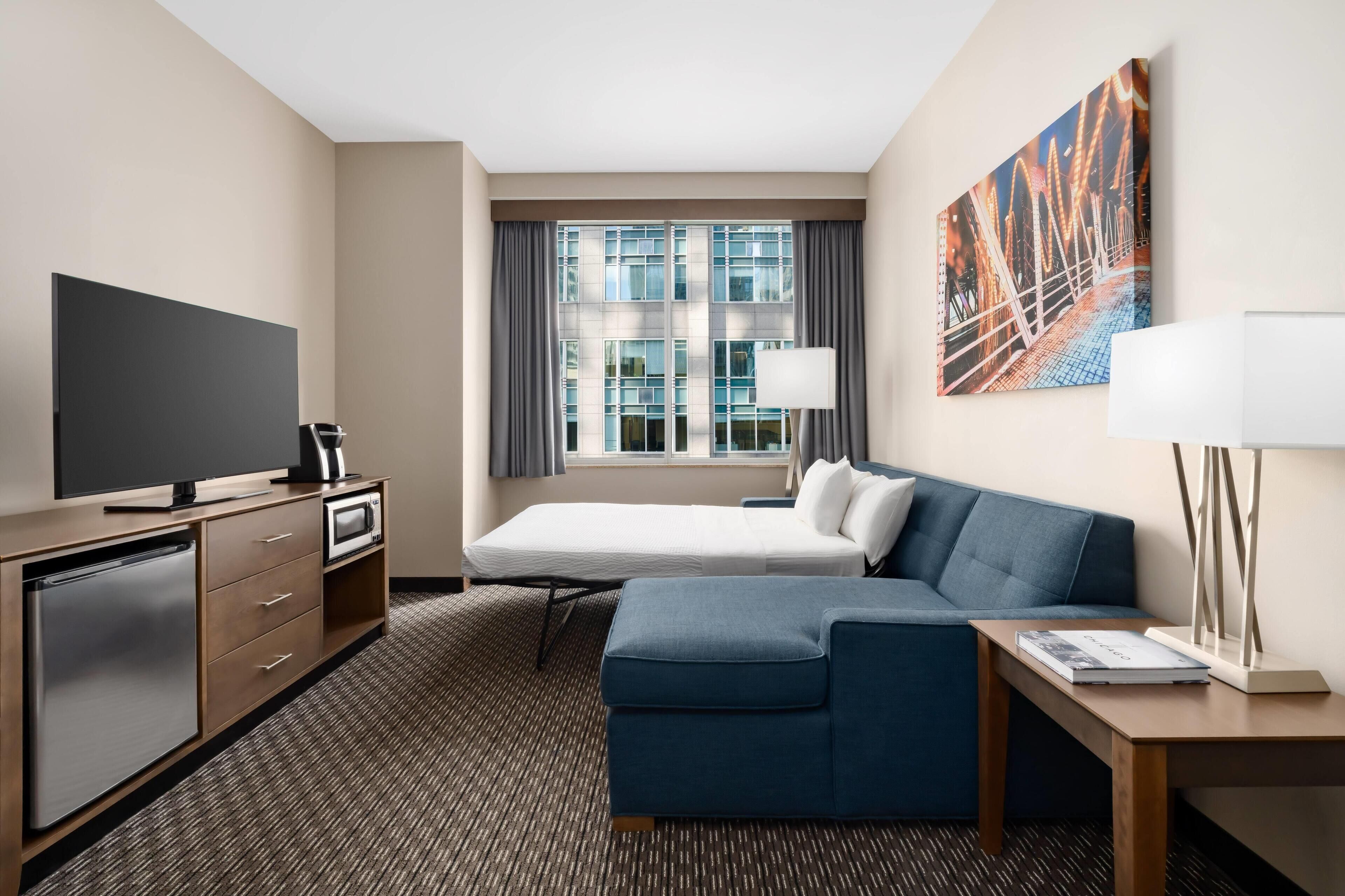 A hotel room with a bed, a sofa, a desk, a TV, a mini-fridge, a lamp, and a painting on the wall.