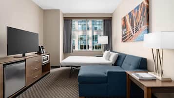 A hotel room with a bed, a sofa, a desk, a TV, a mini-fridge, a lamp, and a painting on the wall.