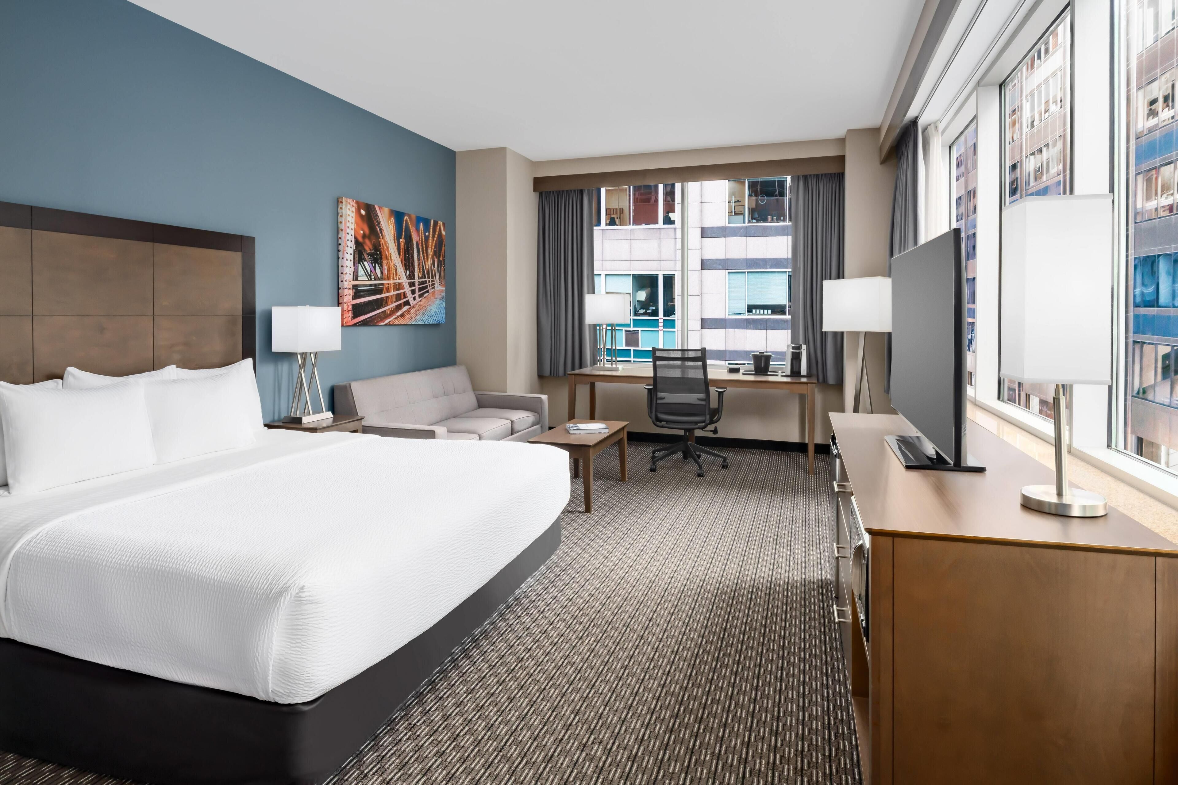 A hotel room with a large bed, a desk, a chair, a TV, and a window with a city view.