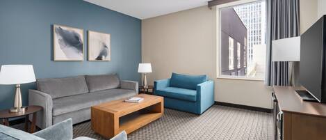 A hotel room with a grey sofa, a blue armchair, a wooden coffee table, and a TV on a wooden cabinet.