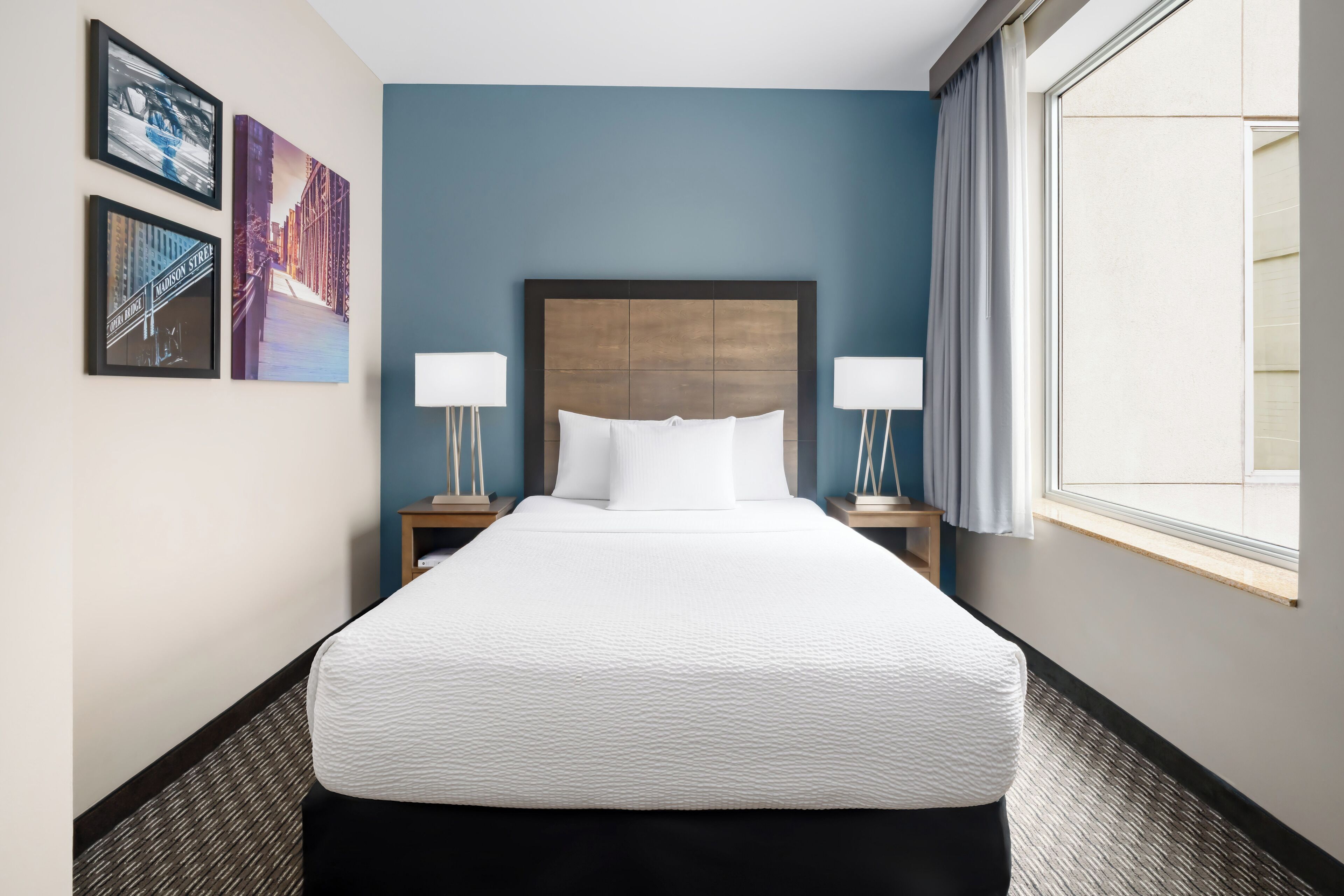 A hotel room with a large bed, two bedside tables with lamps, a window with curtains, and three framed pictures on the wall.