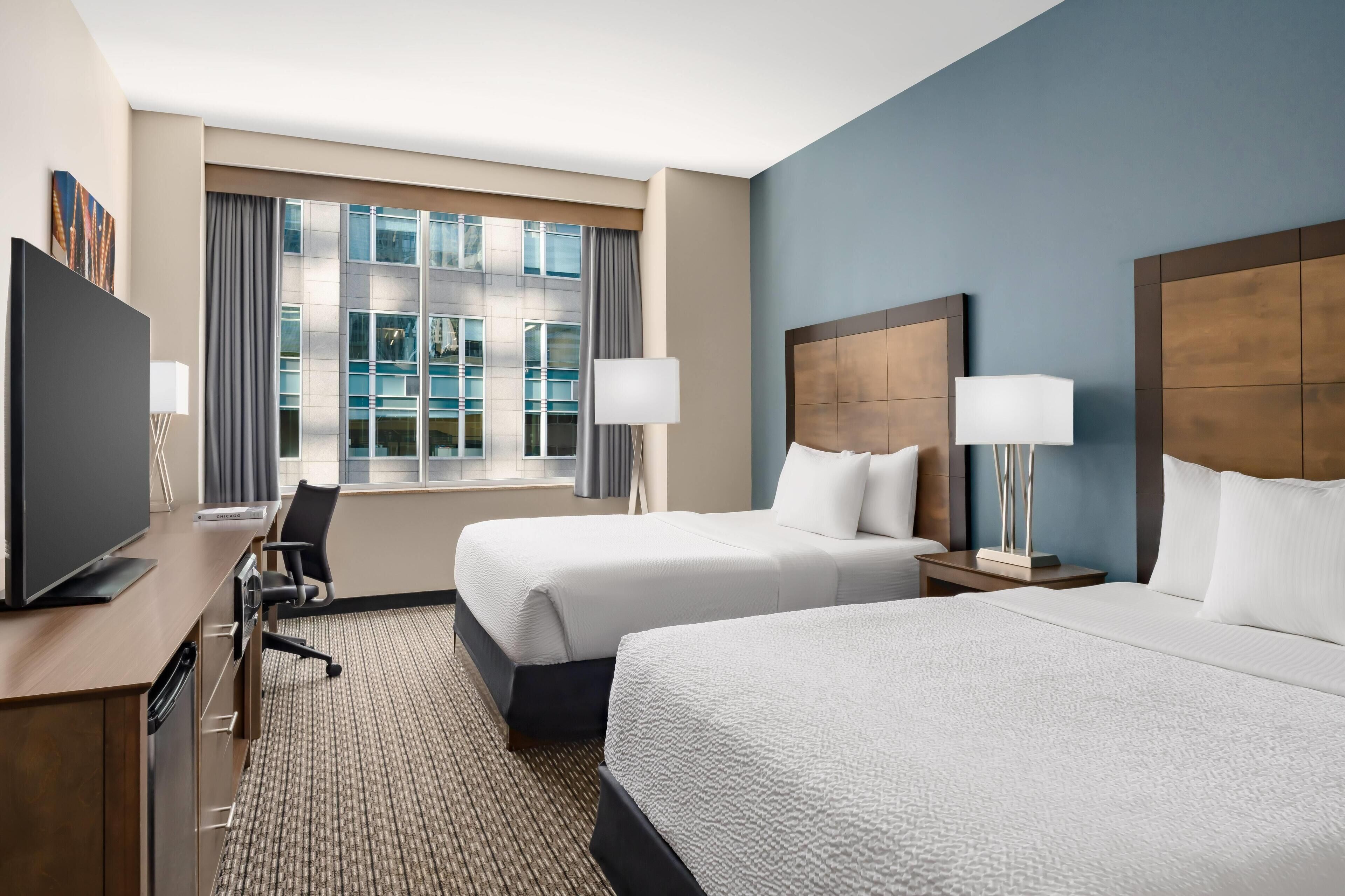 A hotel room with two beds, a desk with a television, and a large window with curtains.