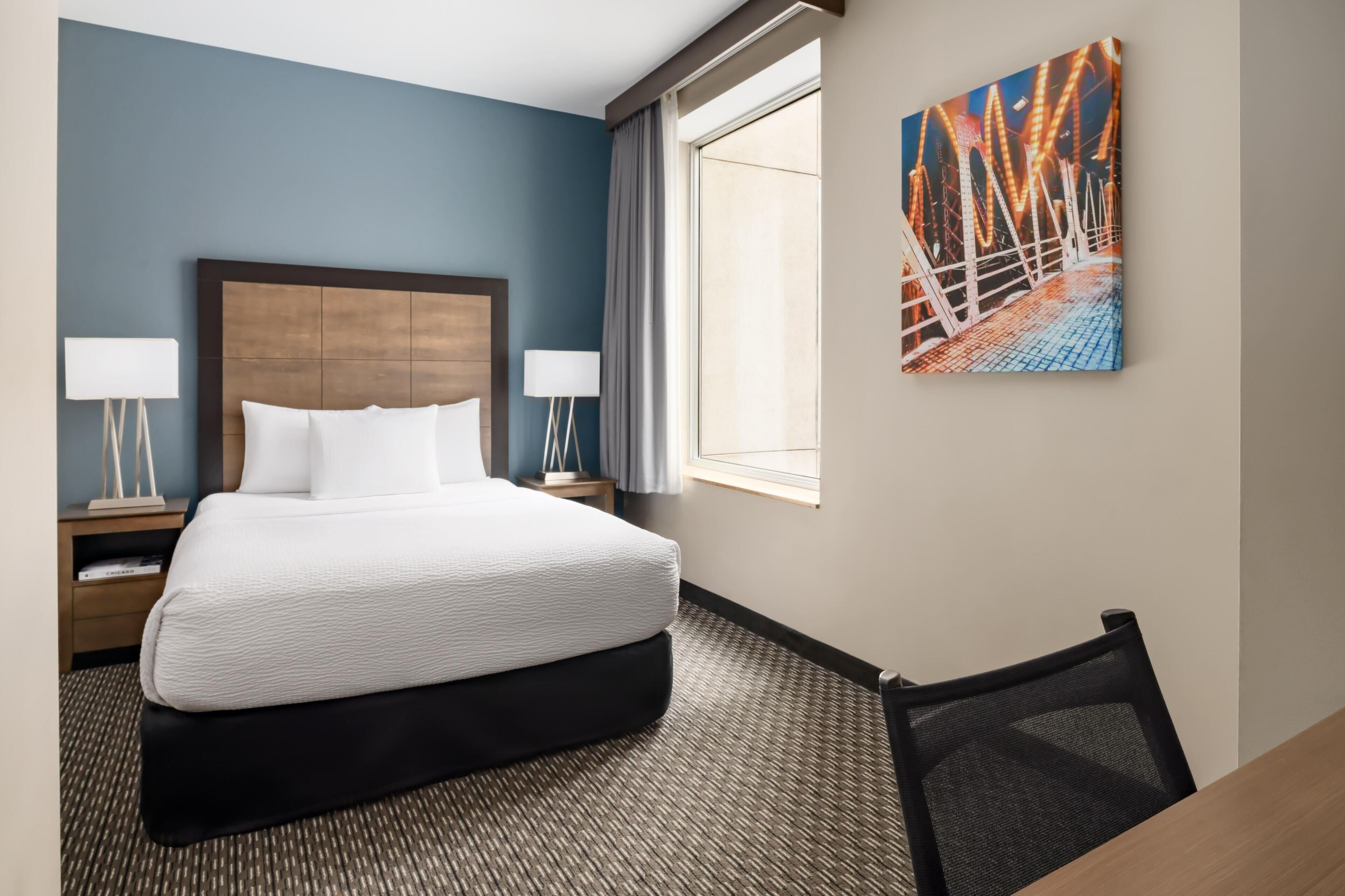 A hotel room with a bed, two bedside tables with lamps, a large window, and a wall-mounted painting.