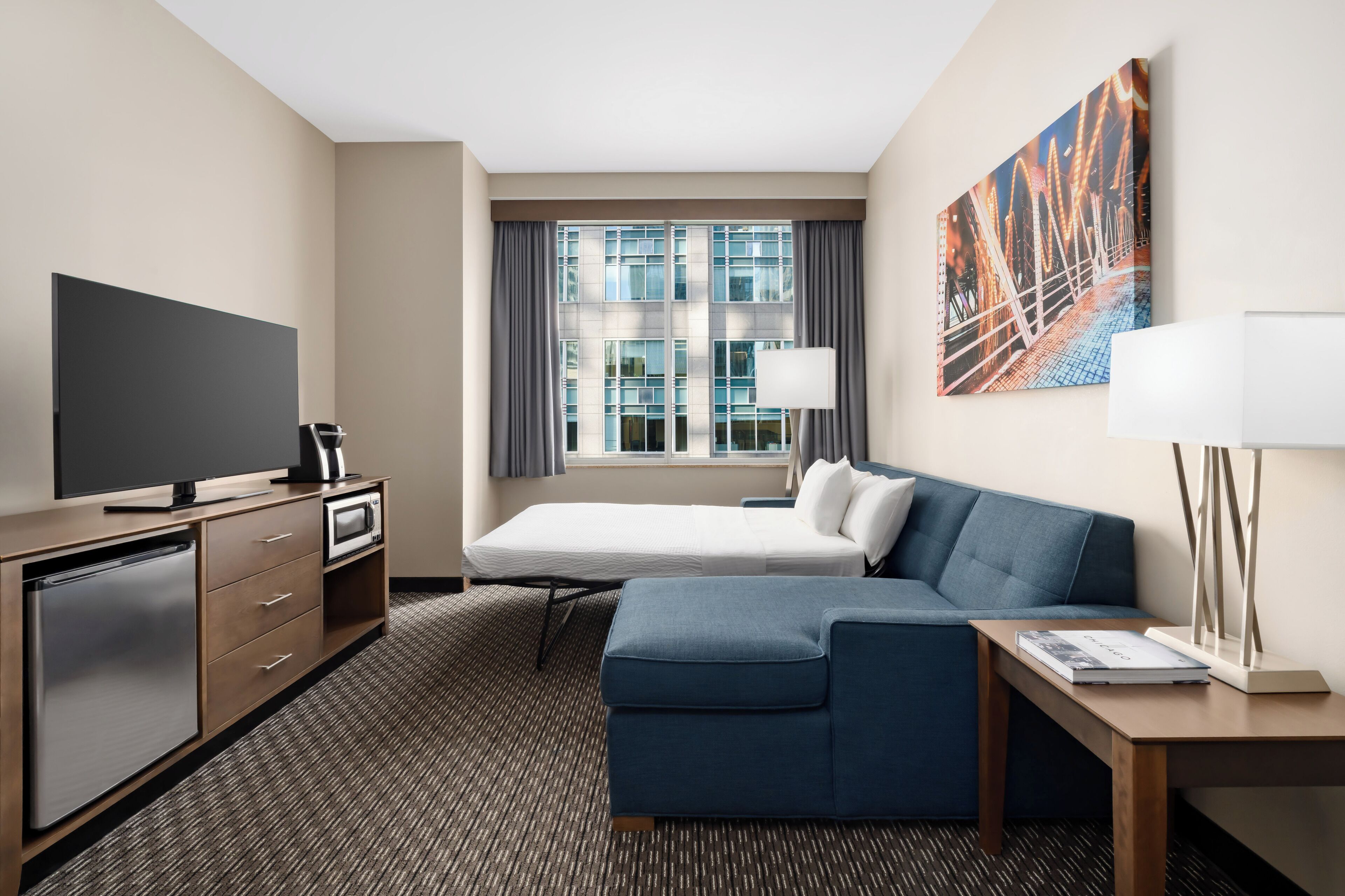 A hotel room with a bed, a TV, a desk, a lamp, and a painting on the wall.