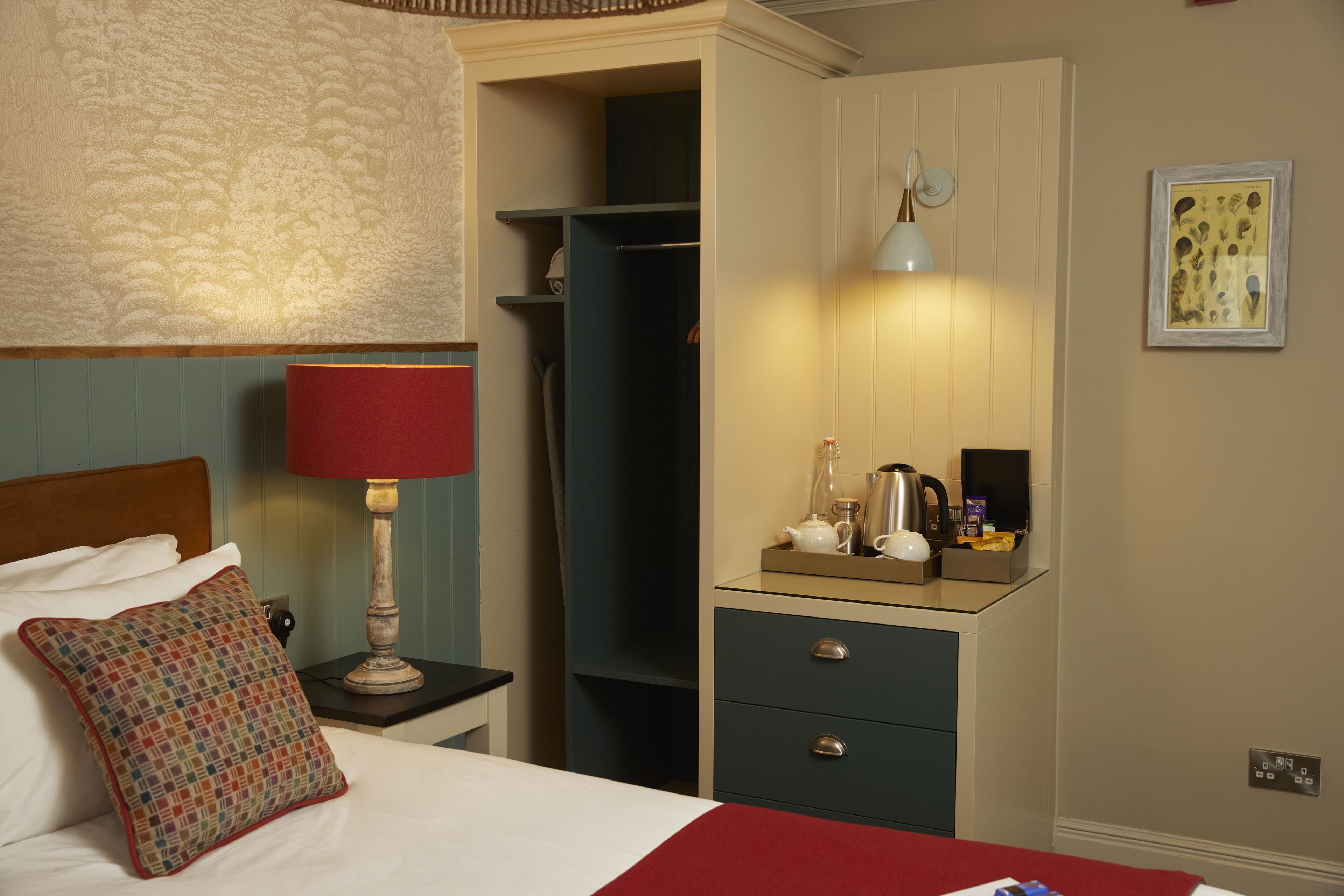A hotel room with a bed, a nightstand with a lamp, a dresser with drawers, a closet, and a small table with a kettle and cups.