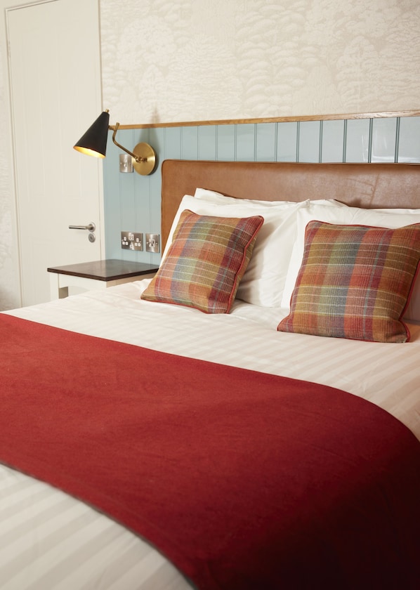 Una cama bien hecha con un edredón rojo, dos almohadas escocesas, un cabecero de madera y una lámpara de noche.