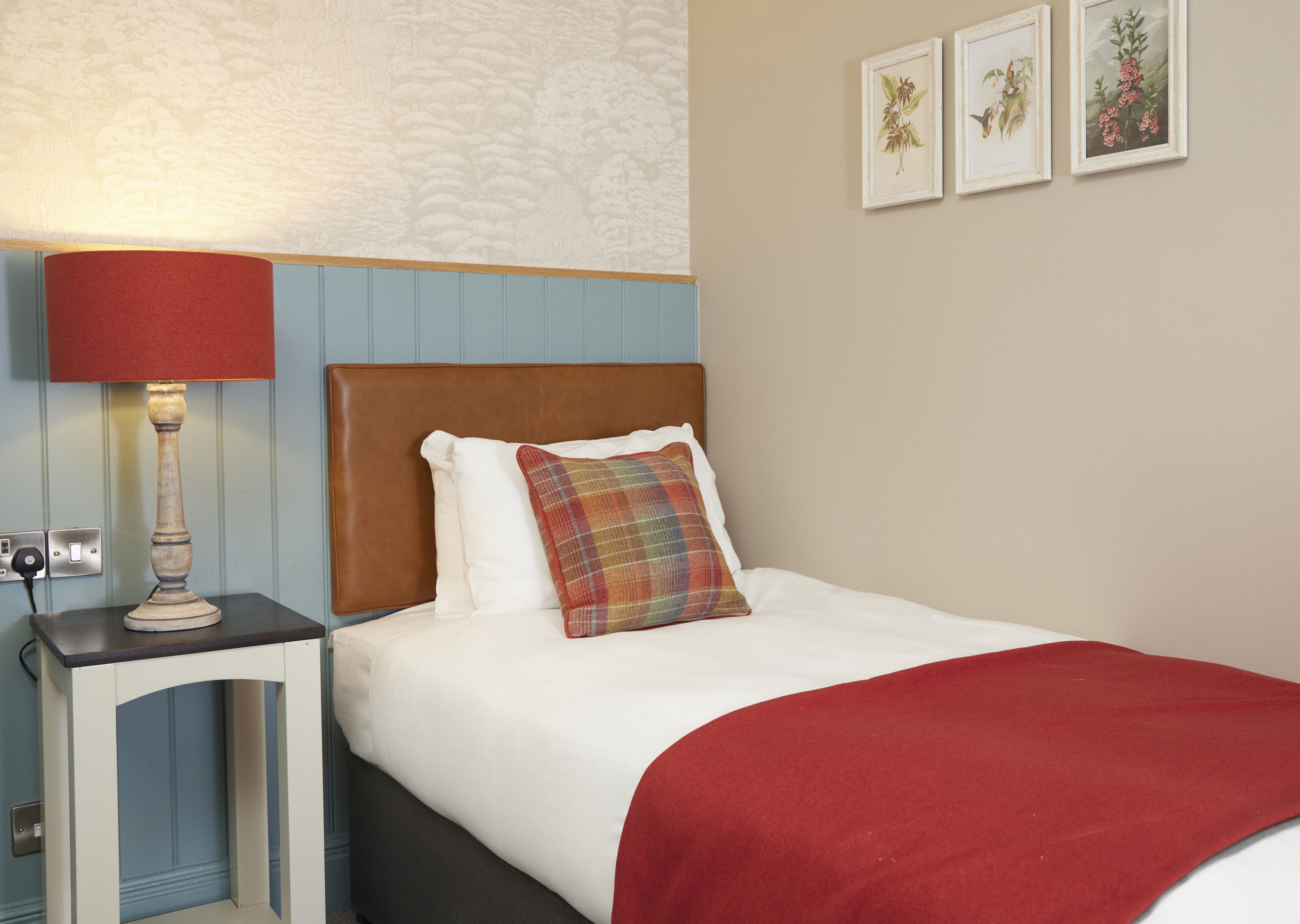 A neatly made bed with a red blanket, a plaid pillow, and a wooden nightstand with a lamp.