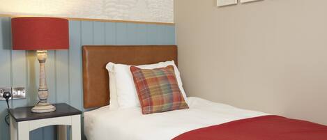 A neatly made bed with a red blanket, a plaid pillow, and a wooden nightstand with a lamp.