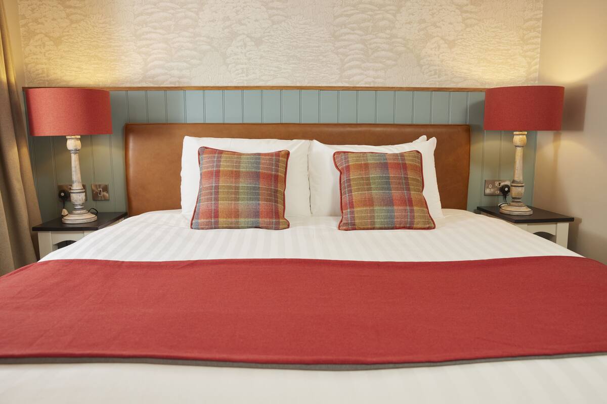 A neatly made bed with a red bedspread, two plaid pillows, and two bedside lamps with red shades.