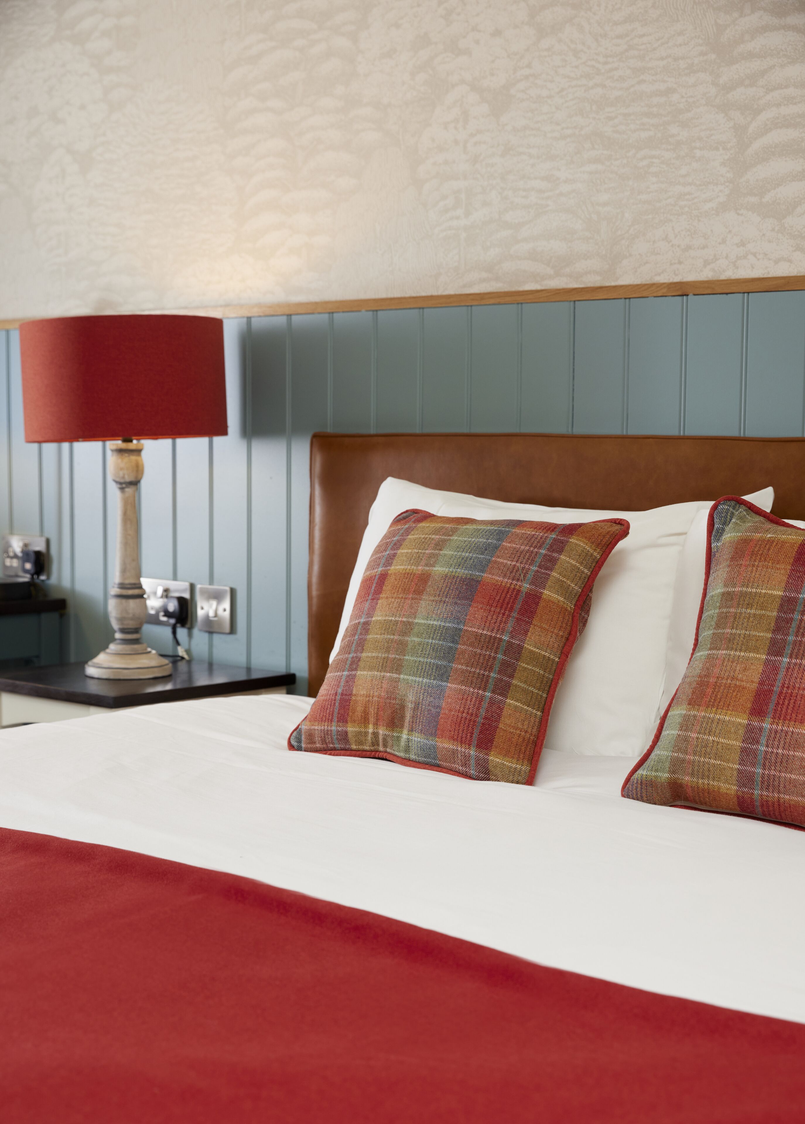 A neatly made bed with plaid pillows and a red blanket, a wooden nightstand with a lamp, and a wall with a subtle floral pattern.