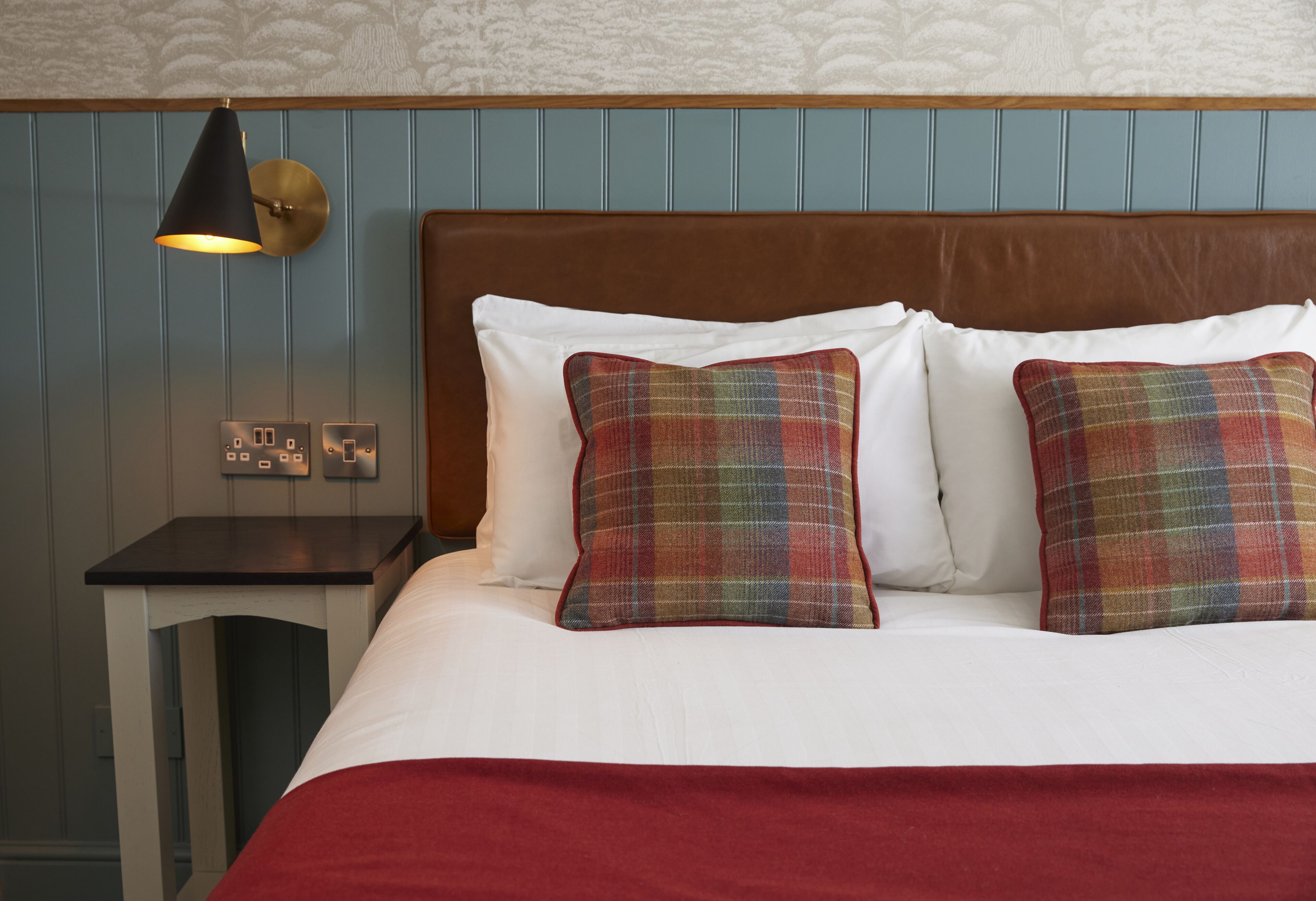 A neatly made bed with white linens and plaid pillows, a wooden headboard, and a bedside table with a lamp.