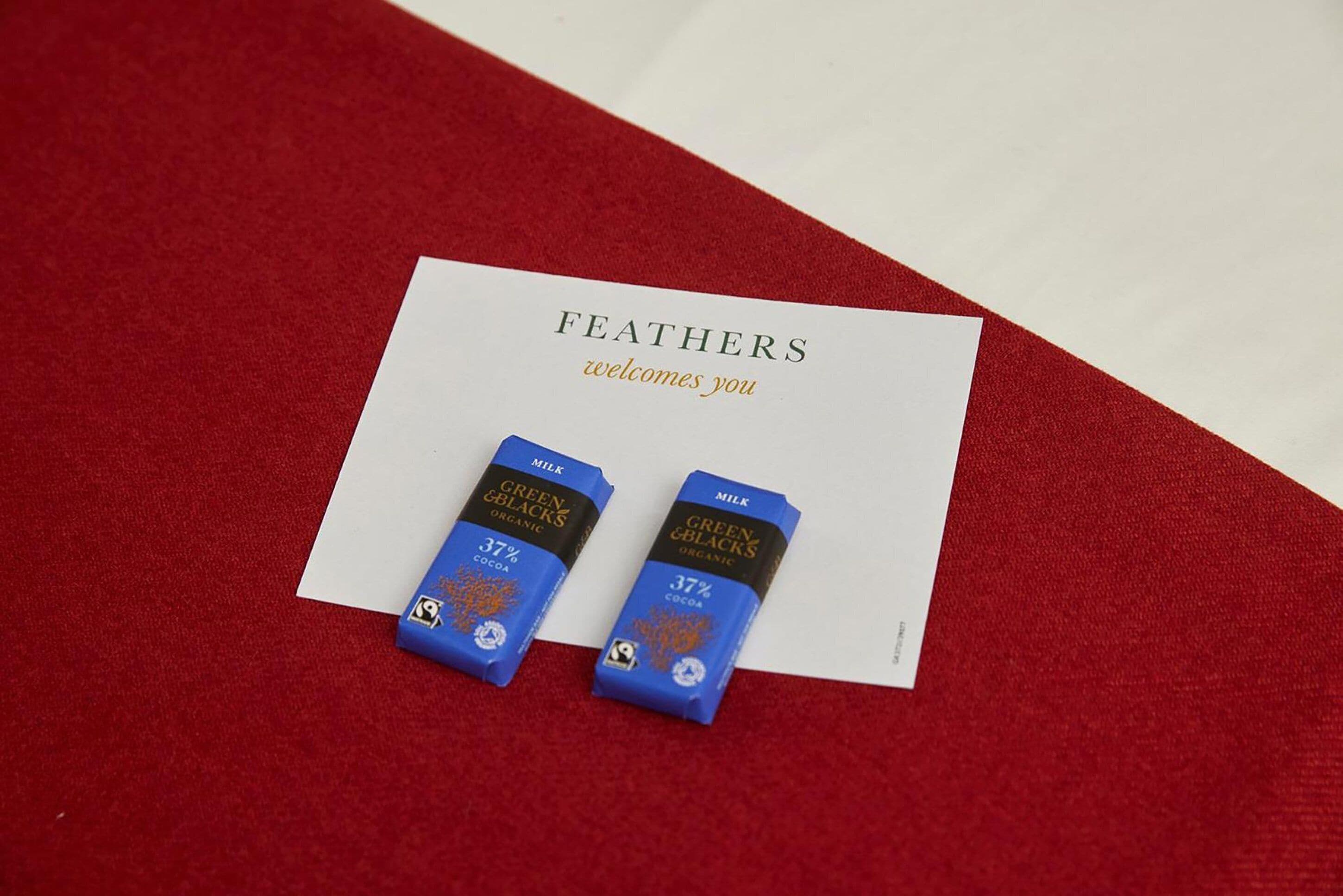 A card with the text "FEATHERS welcomes you" and two small blue boxes labeled "GREEN BLACKS ORGANIC MILK 37% COCOA".