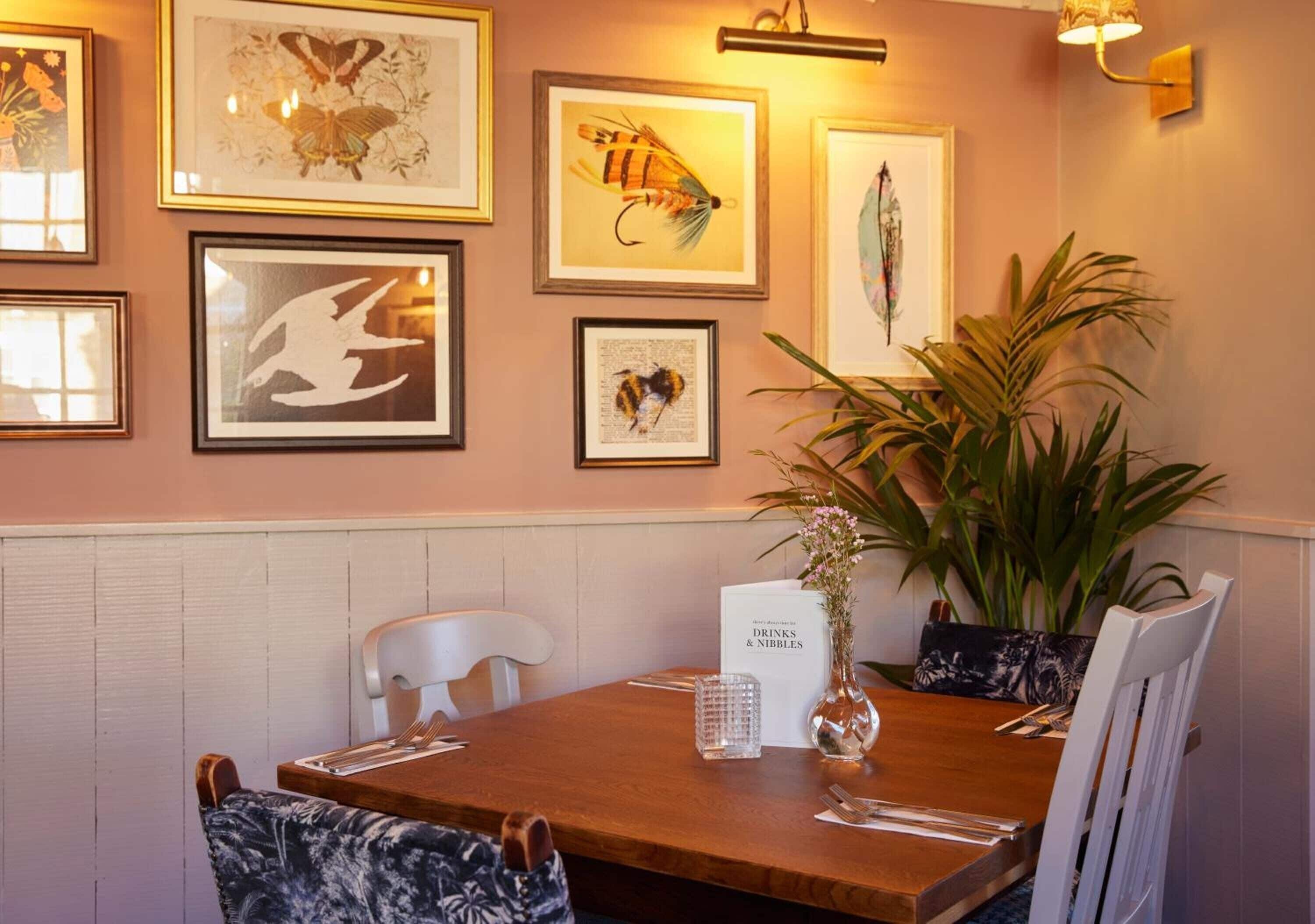 Un comedor con una mesa de madera, sillas blancas, una obra de arte enmarcada en la pared, un atril para el menú y una planta en maceta.