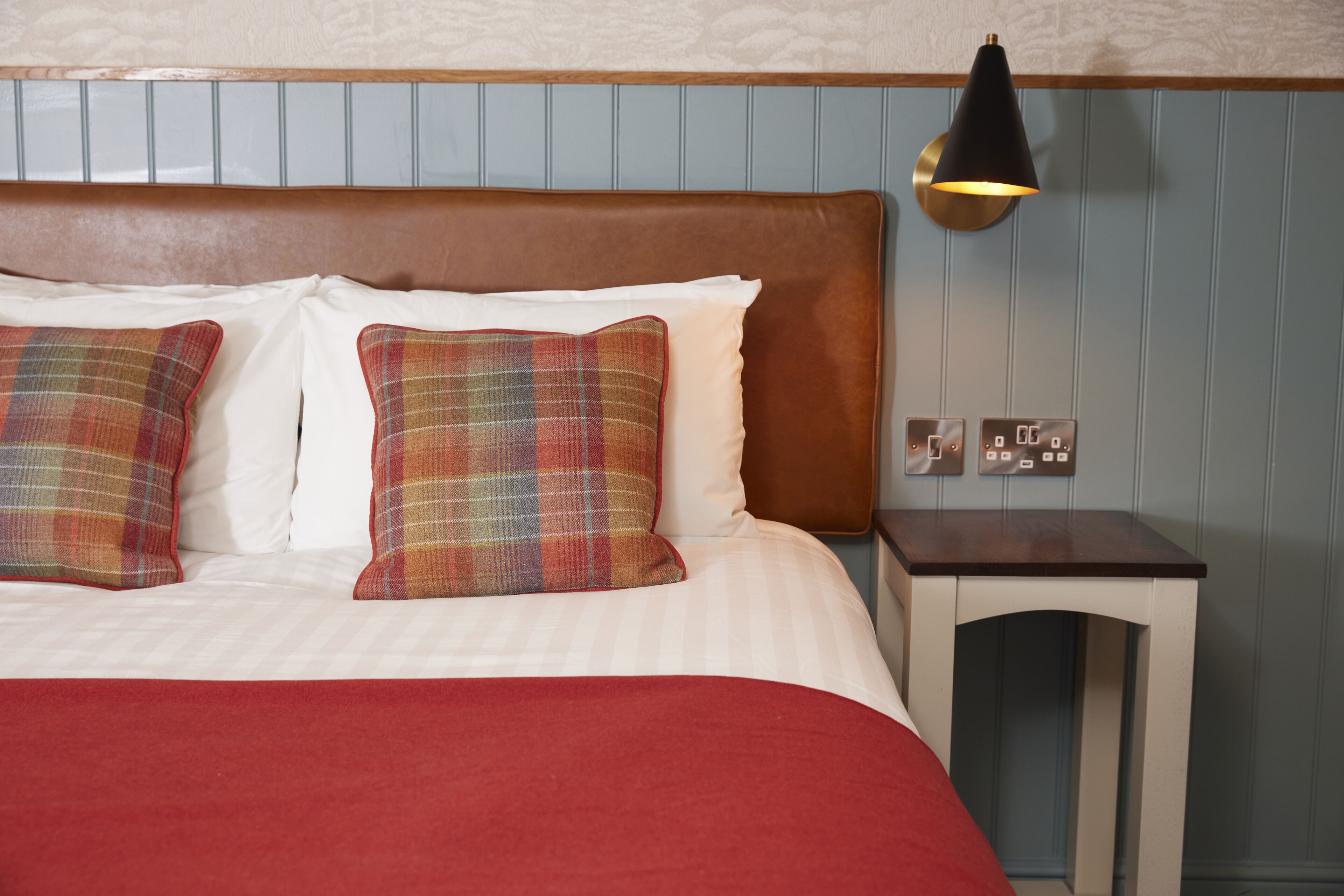 A neatly made bed with plaid pillows, a red blanket, and a bedside table with a lamp.