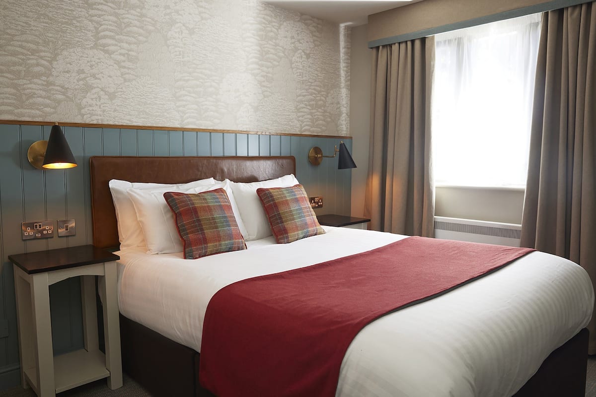 A neatly made bed with a red blanket, two plaid pillows, and a wooden headboard. There are two bedside tables with lamps, and a window with curtains.