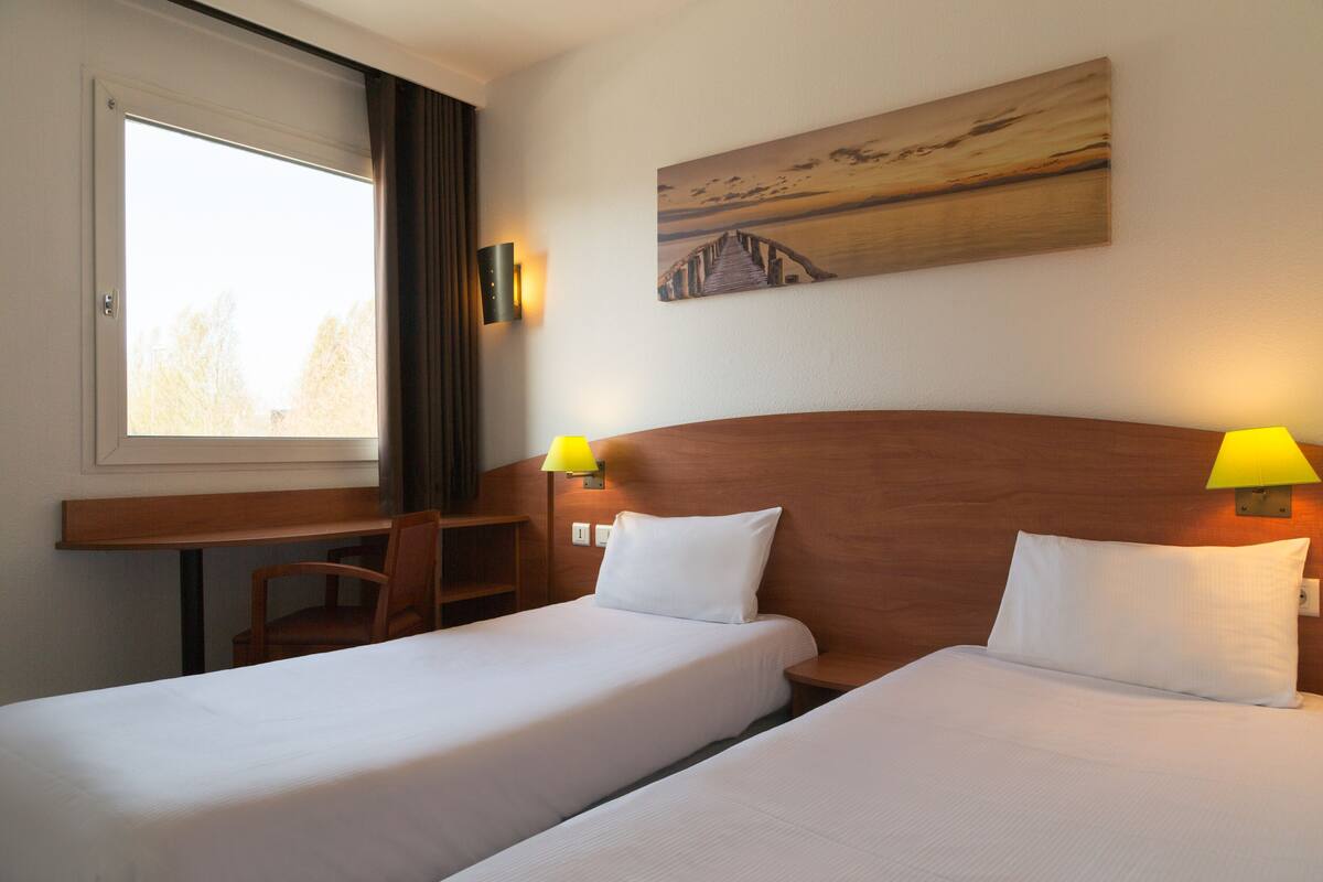 Hotelkamer met twee bedden, een bureau en een stoel. Er hangt een schilderij aan de muur en er is een raam met gordijnen.