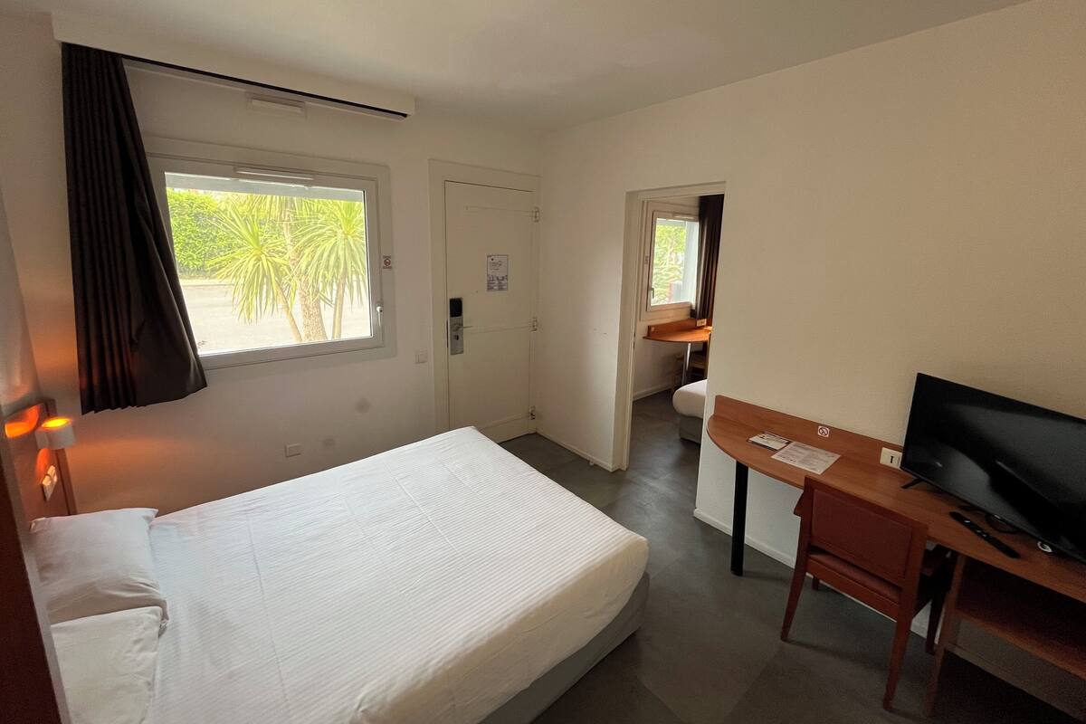 Een hotelkamer met een bed, een bureau met televisie, een raam met gordijnen en een deur die naar een andere kamer leidt.