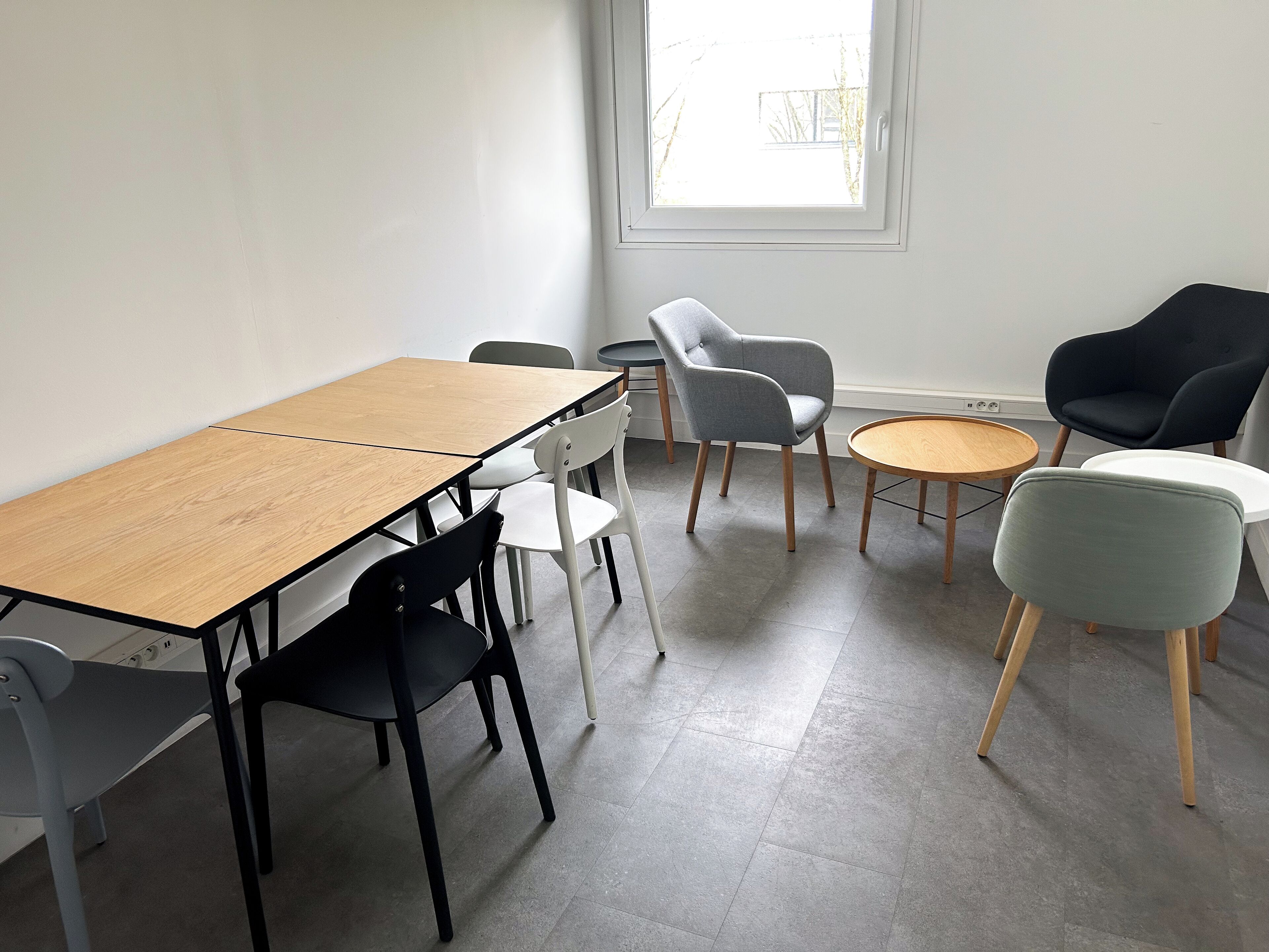 Una habitación con una mesa de madera, sillas blancas y negras, un sillón gris y un sillón negro, además de una mesa redonda de madera.