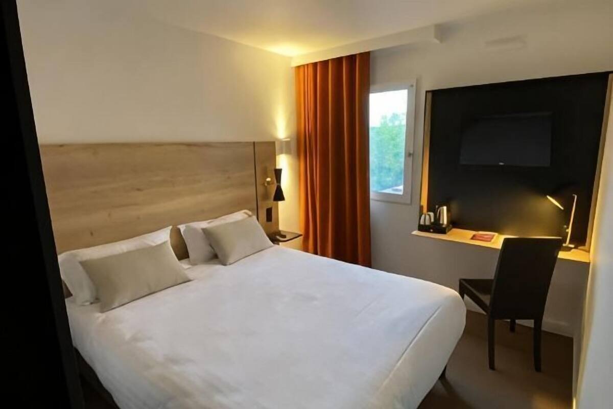 A hotel room with a bed, a desk with a chair, a TV, and a window with curtains.