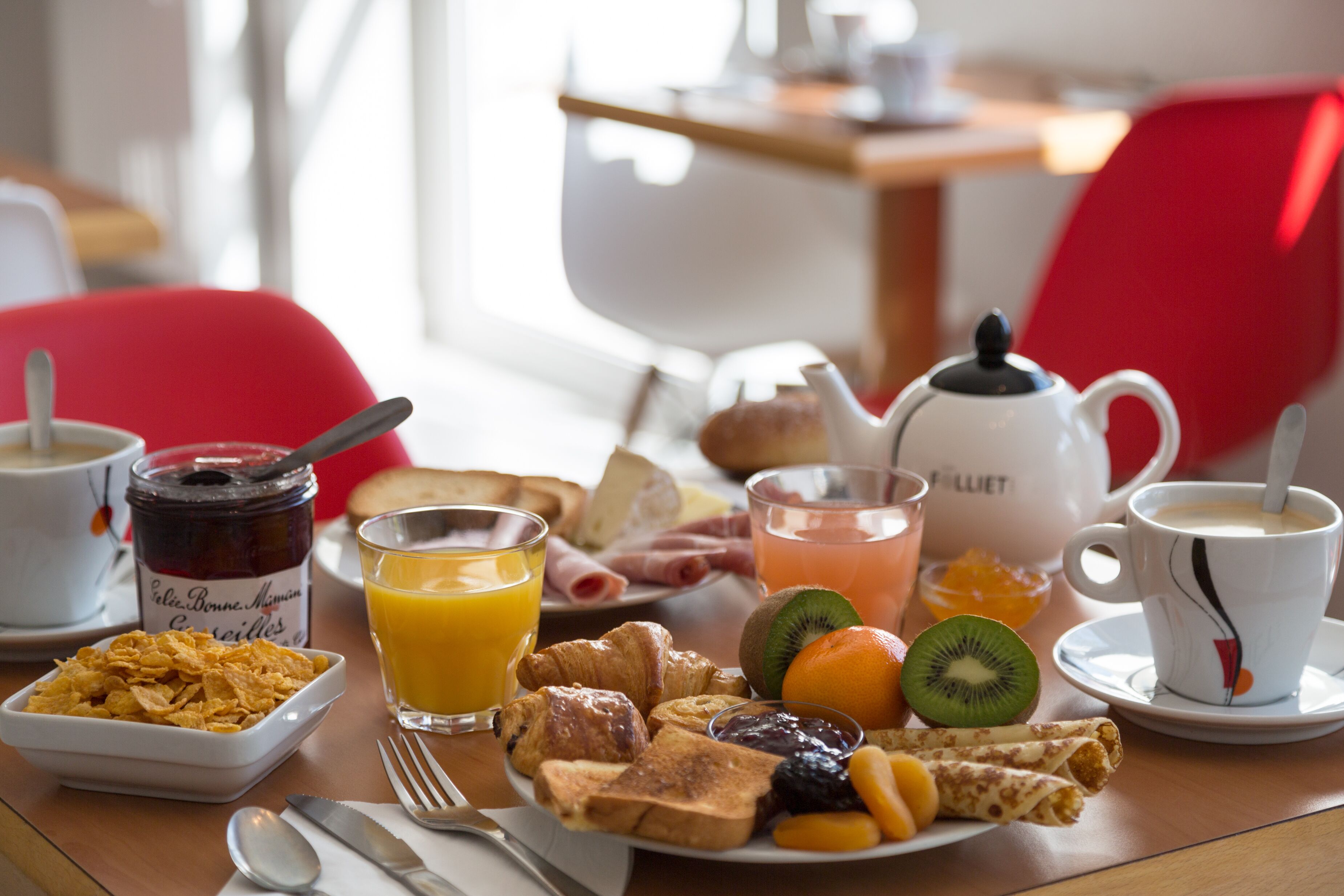 A breakfast table with cereal, juice, croissants, fruits, and a teapot.