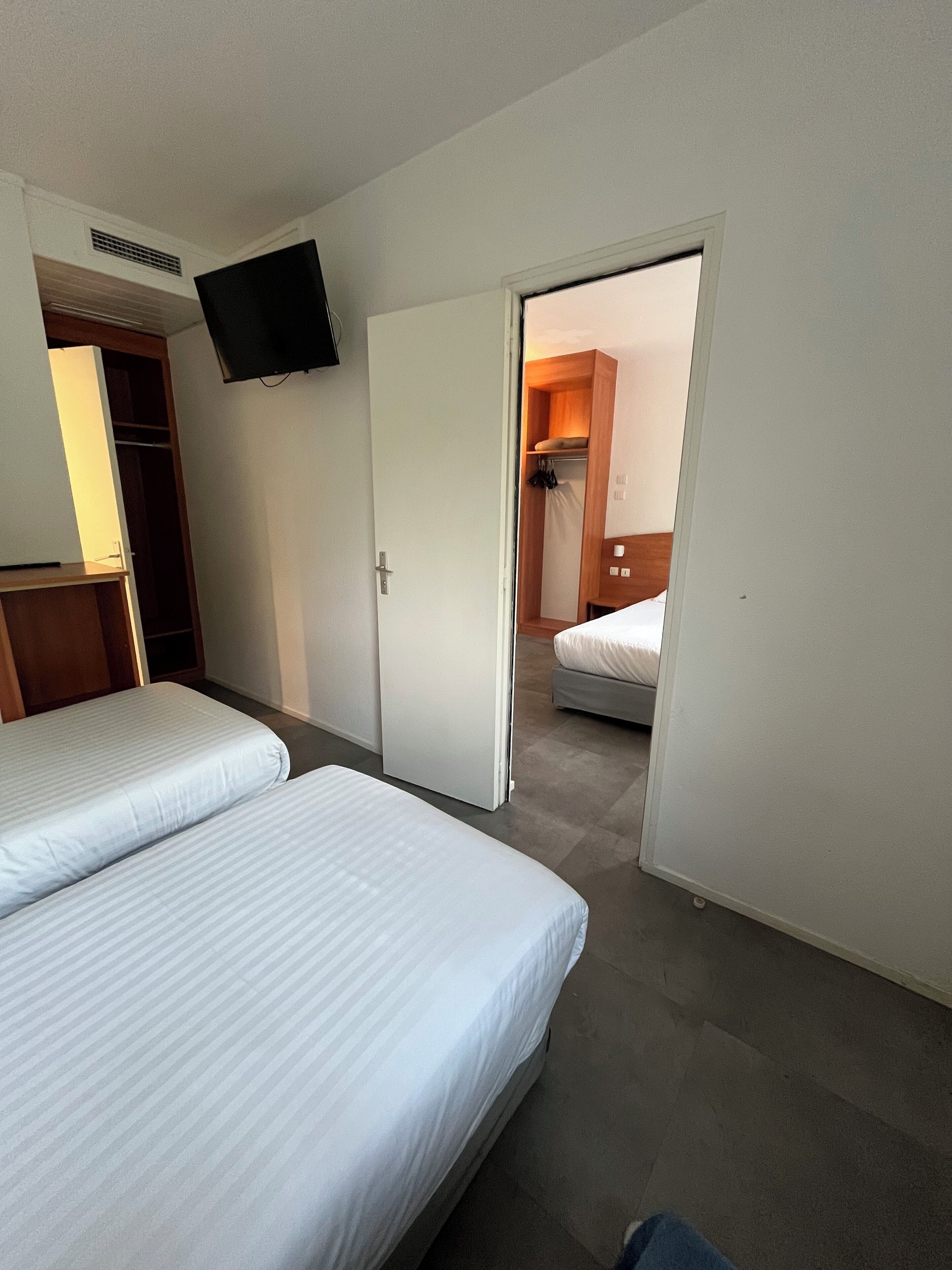 A hotel room with two beds, a wardrobe, a television mounted on the wall, and a bathroom visible through an open door.