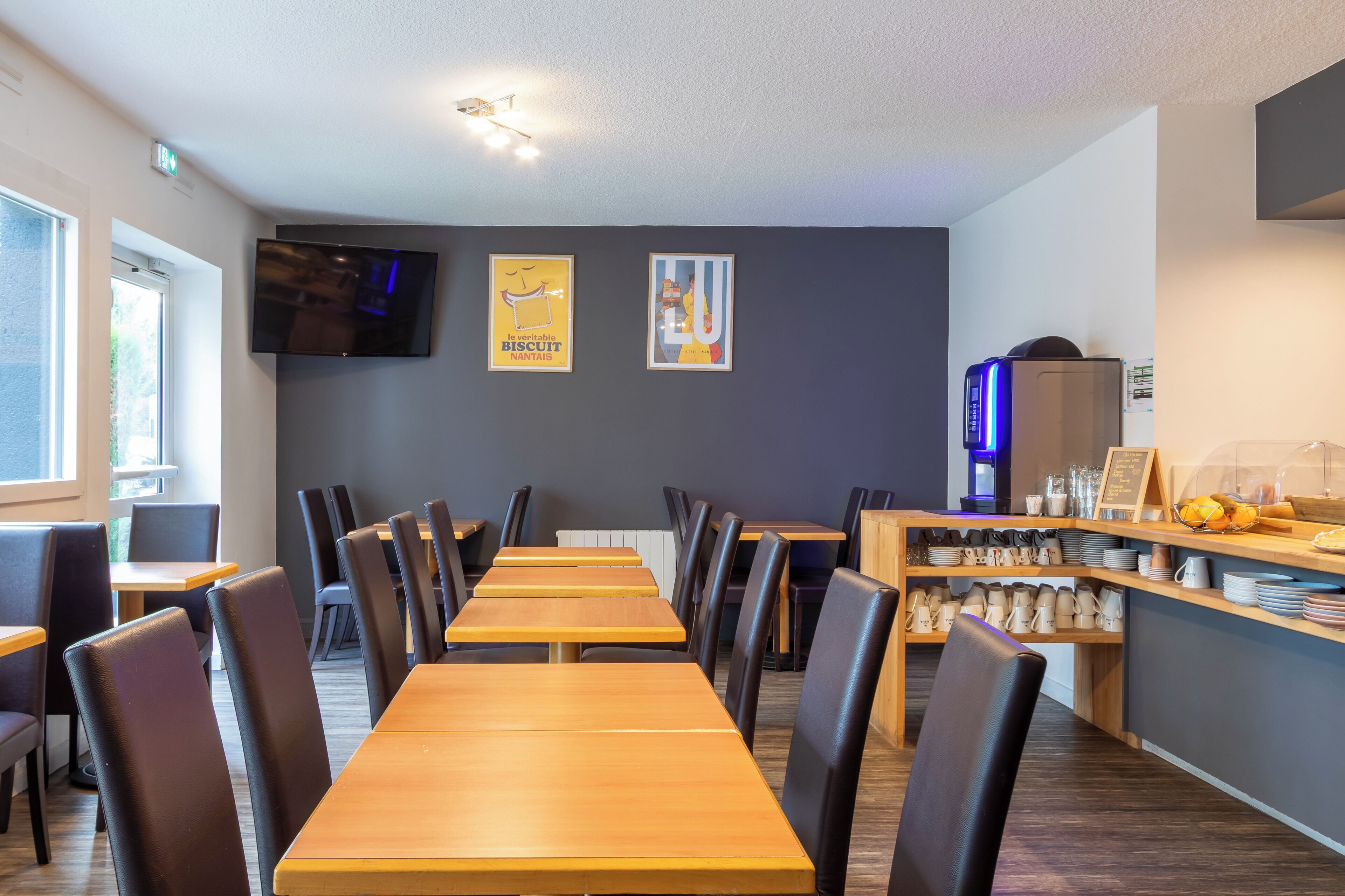Een restaurant met lange houten tafels en zwarte stoelen, een televisie aan de muur en een toonbank met diverse artikelen.