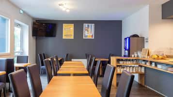 Een restaurant met lange houten tafels en zwarte stoelen, een televisie aan de muur en een toonbank met diverse artikelen.