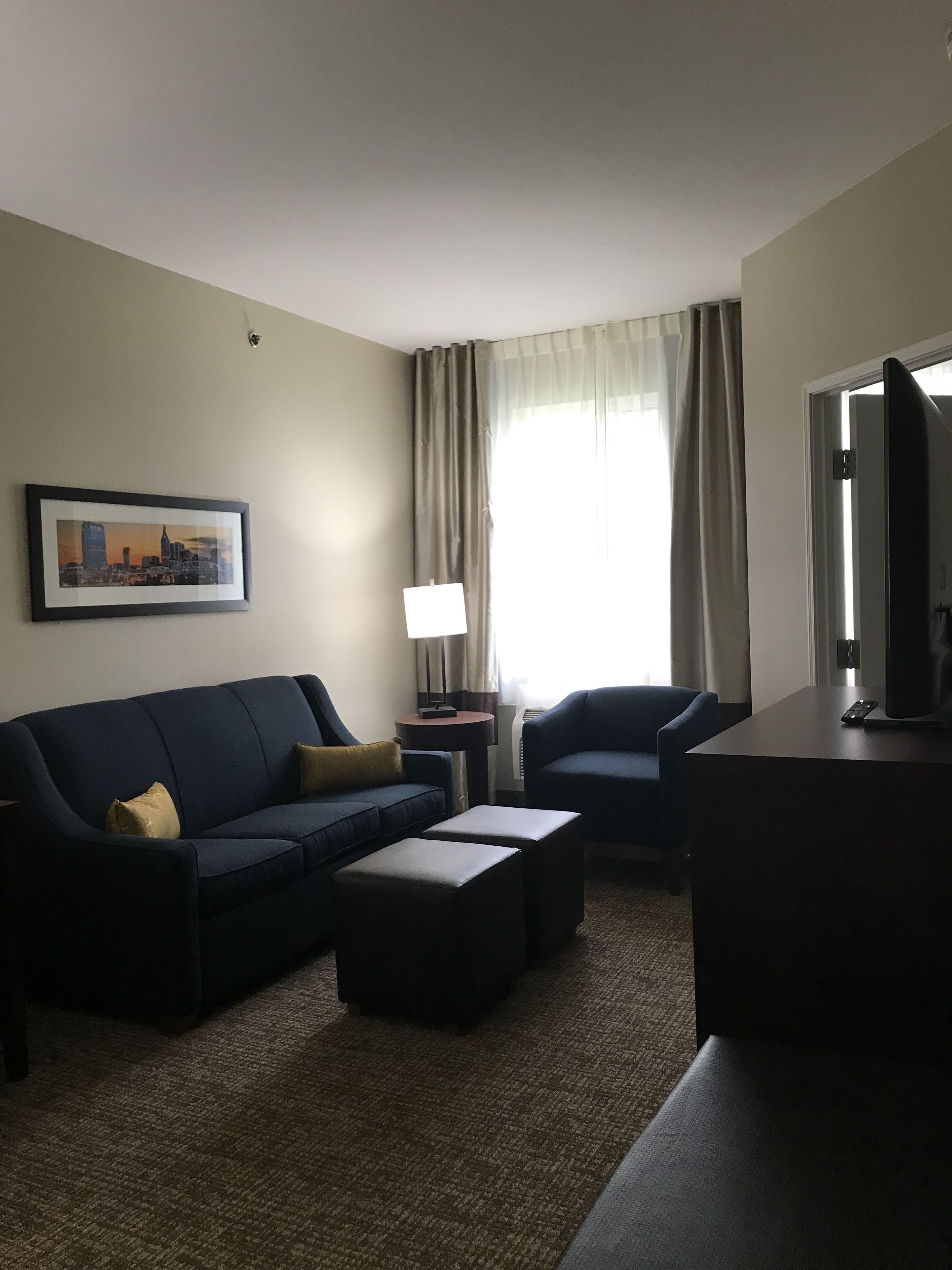 A modern hotel room with a dark blue sofa, a matching armchair, a small side table with a lamp, and a flat-screen TV on a stand.