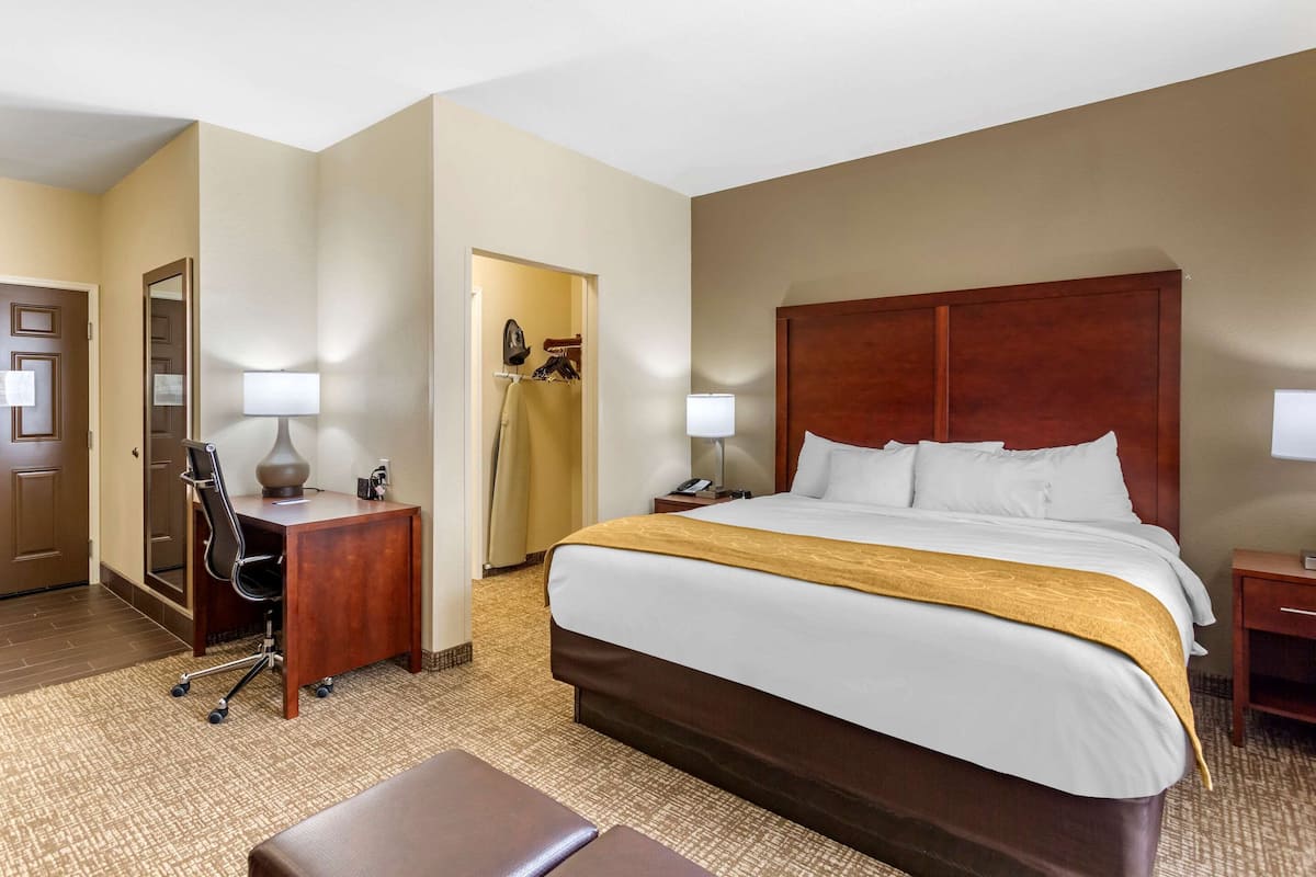 A hotel room with a large bed, a desk with a chair, a desk lamp, a nightstand, and a wardrobe.