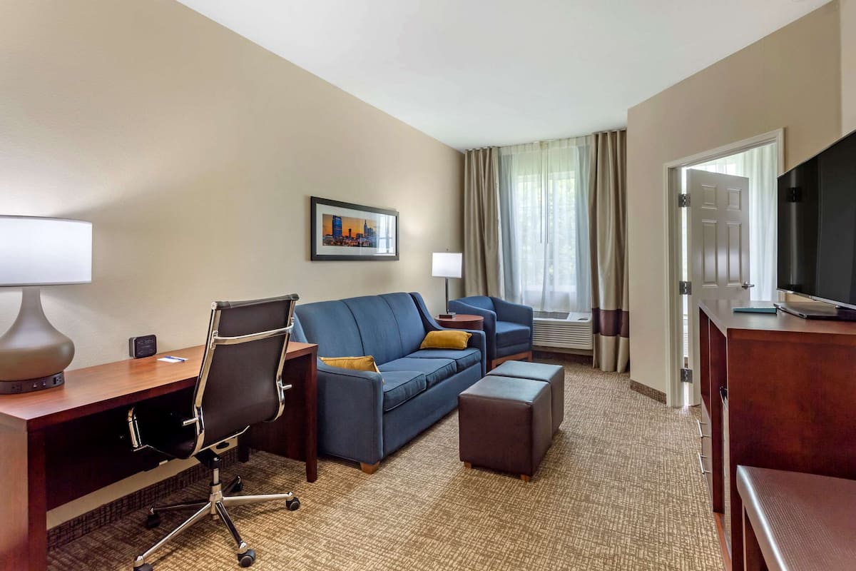 A hotel room with a desk, two sofas, a TV, and a window with curtains.
