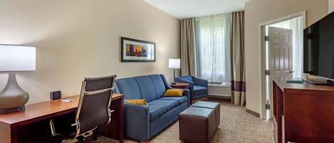 A hotel room with a desk, two sofas, a TV, and a window with curtains.