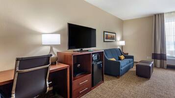 A hotel room with a desk, chair, television, sofa, and a small refrigerator.