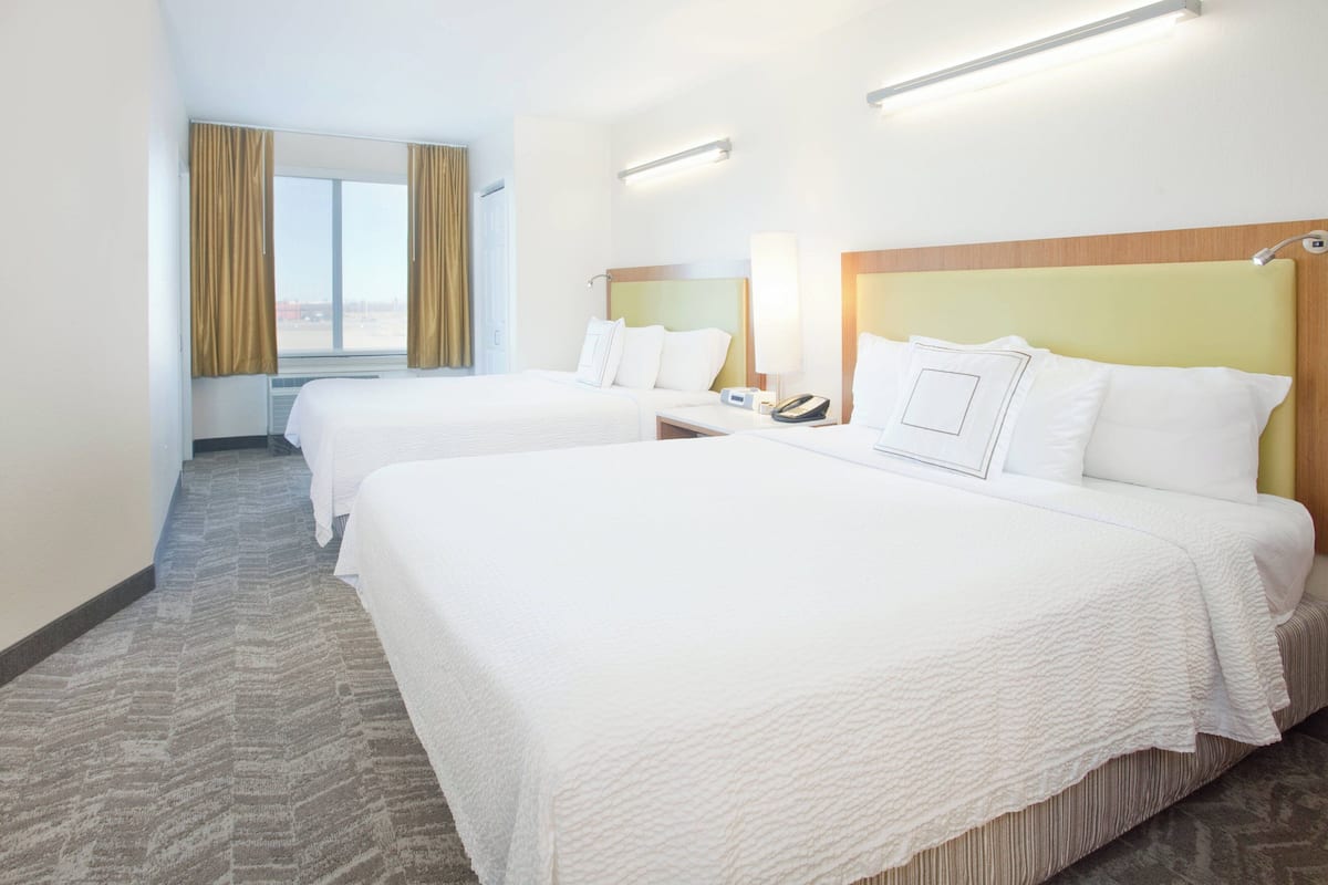 A hotel room with two beds, a window with curtains, a bedside table with a lamp, and a wall-mounted phone.