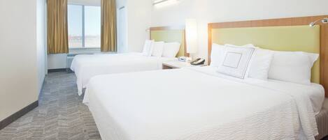 Quarto de hotel com duas camas, uma janela com cortinas, uma mesa de cabeceira com abajur e telefone fixado na parede.